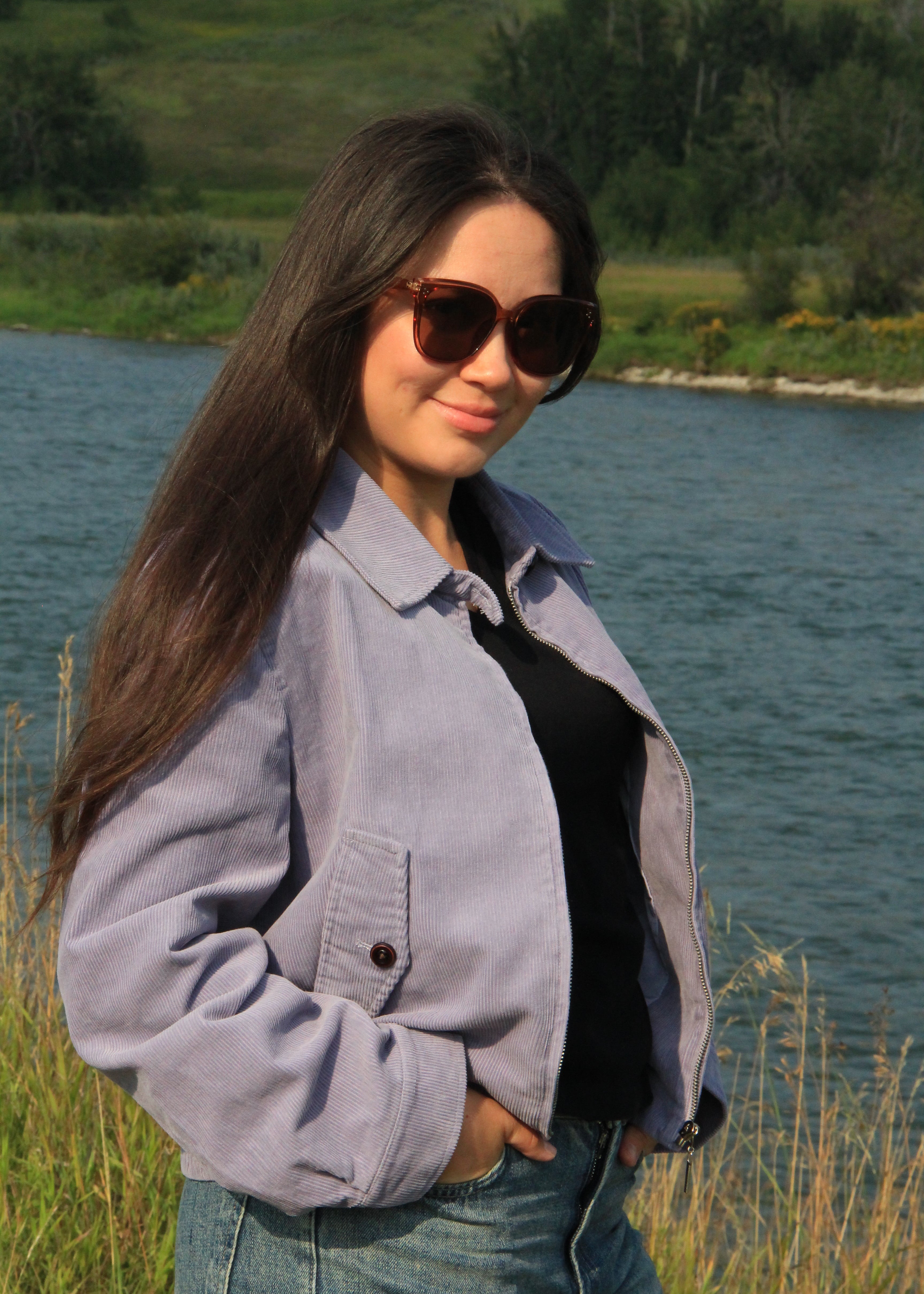 A woman with long dark hair wearing a light purple corduroy jacket over a black top and blue jeans, standing outdoors near a river with grass and trees in the background.