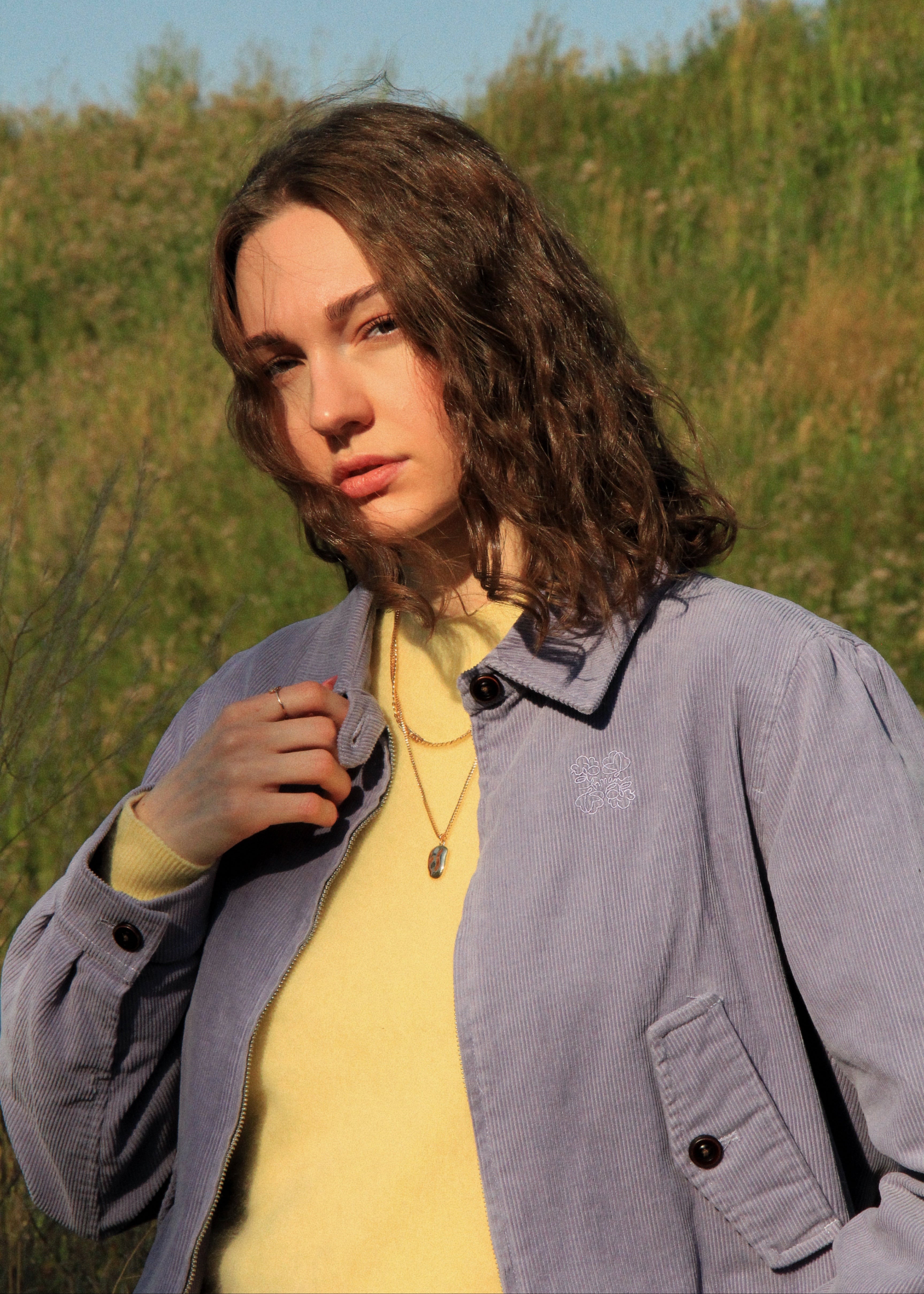 A woman outdoors wearing a light purple-gray overshirt over a pale yellow top, with a gold necklace, against a blurred green and blue background.