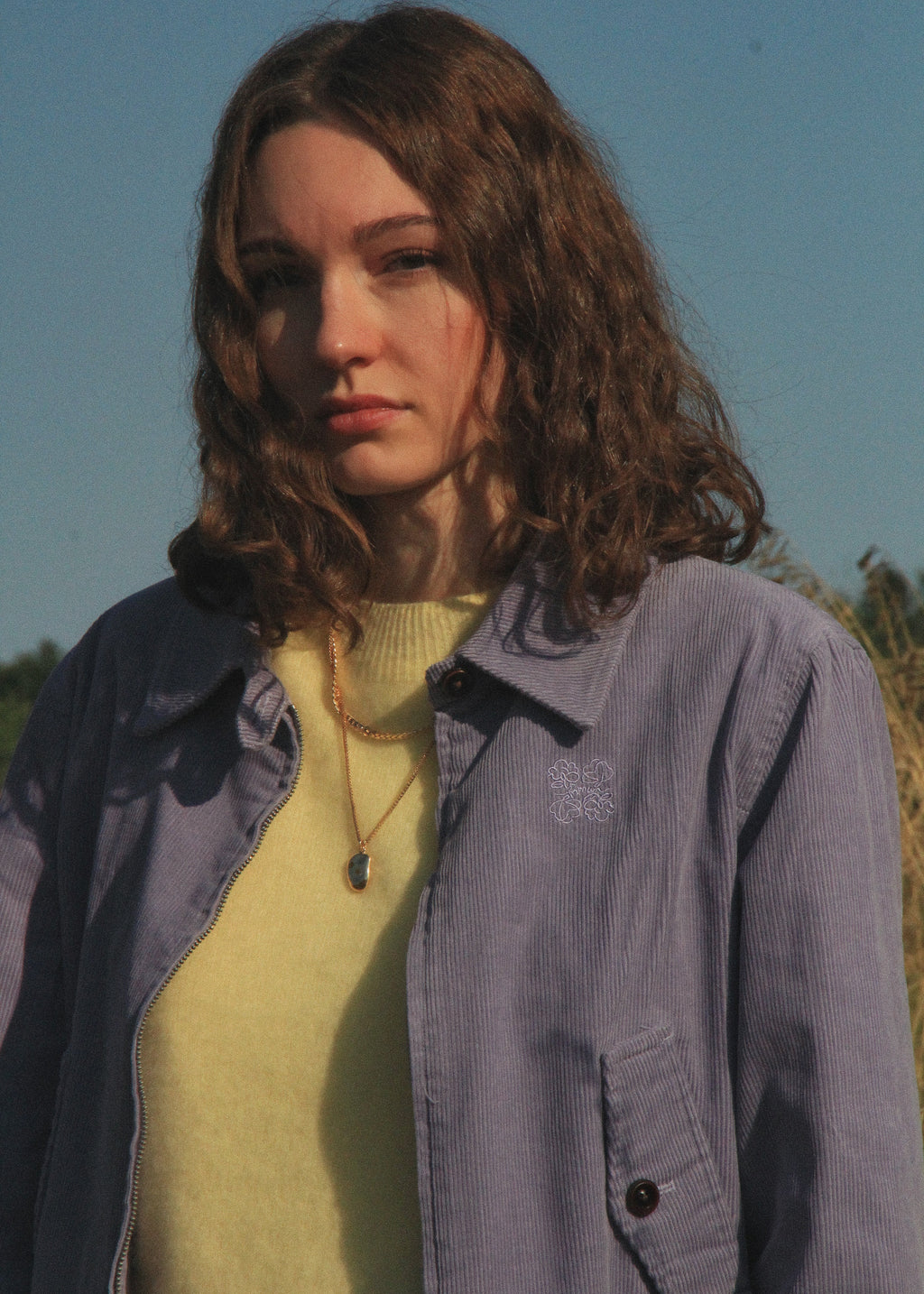 A woman outdoors wearing a light purple corduroy overshirt over a pale yellow knit top, with a small embroidered logo on the chest pocket. The background shows a clear blue sky and some dry grass.