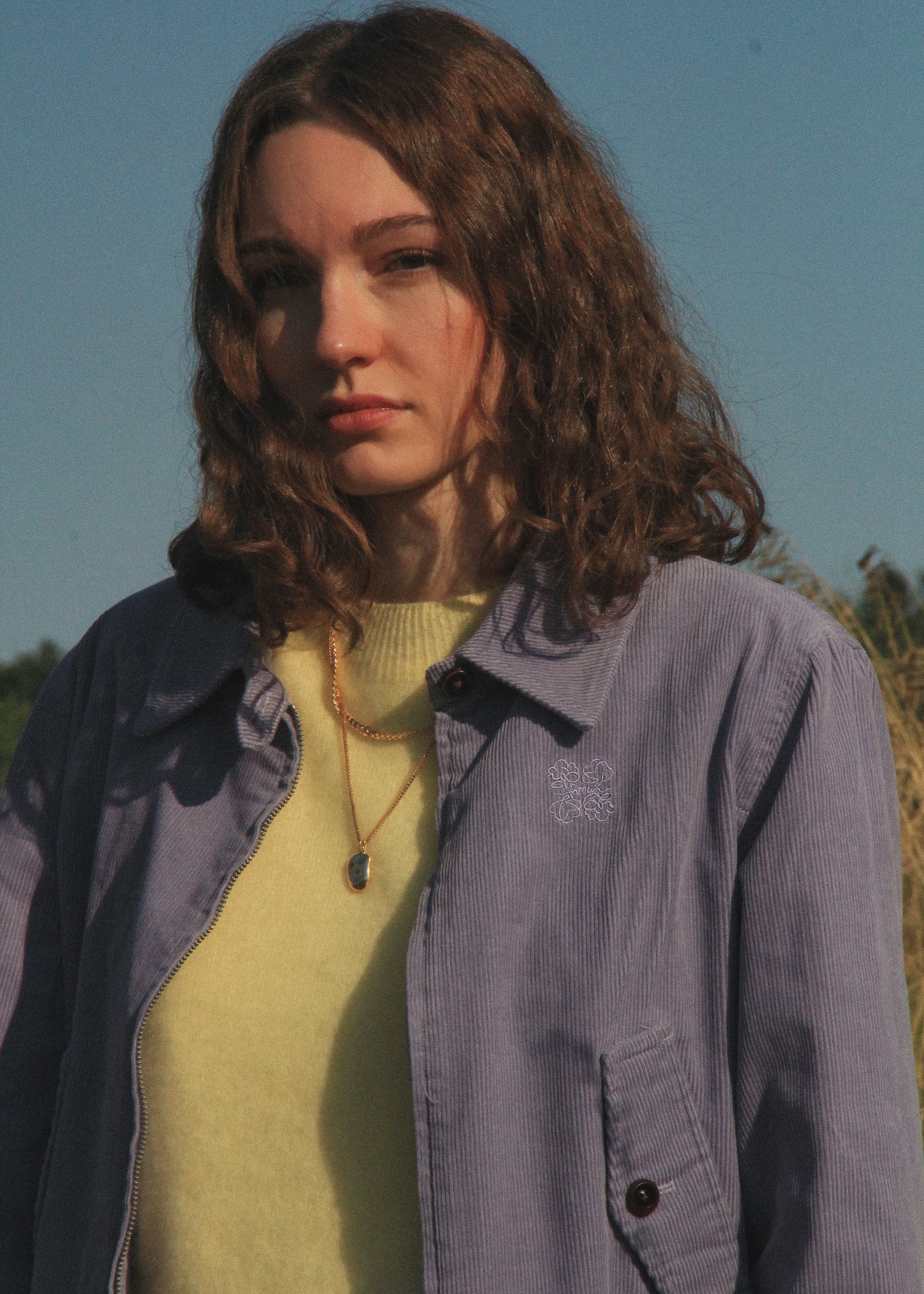 A woman outdoors wearing a light purple corduroy overshirt over a pale yellow knit top, with a small embroidered logo on the chest pocket. The background shows a clear blue sky and some dry grass.