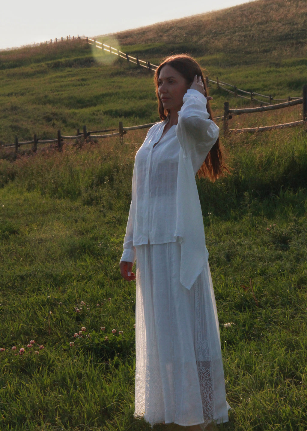 A woman wearing a long white lace dress and matching cardigan stands in a grassy field with a wooden fence and rolling hills in the background during golden hour.