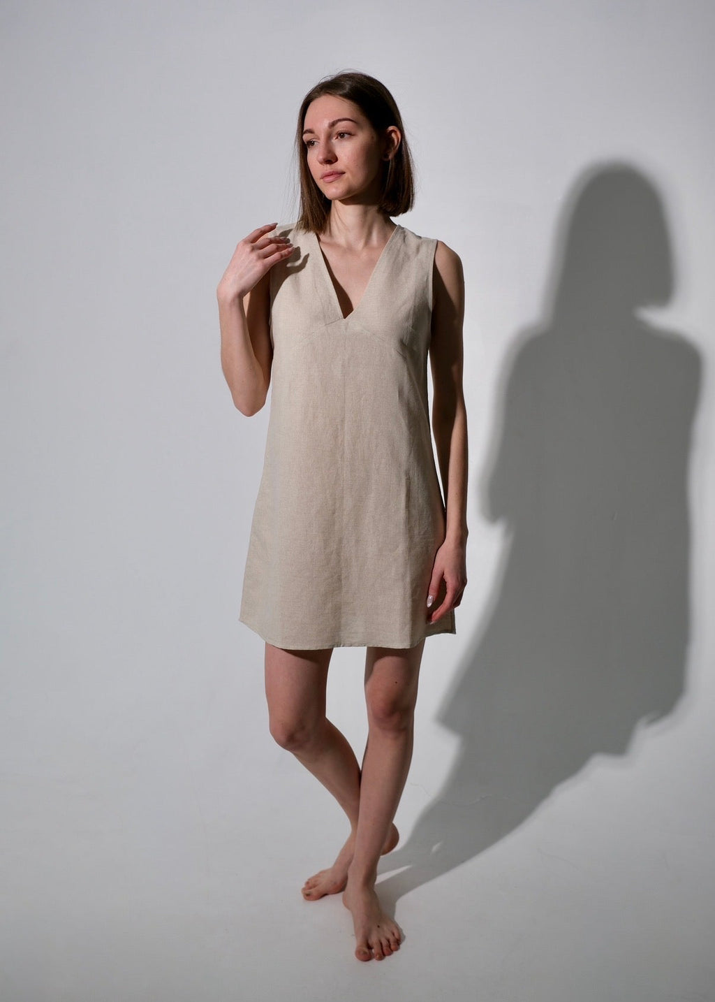 Woman wearing a beige sleeveless dress standing against a white wall with a soft shadow.