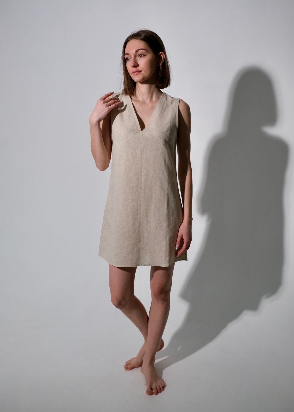 Woman wearing a beige sleeveless dress standing against a white wall with a soft shadow.