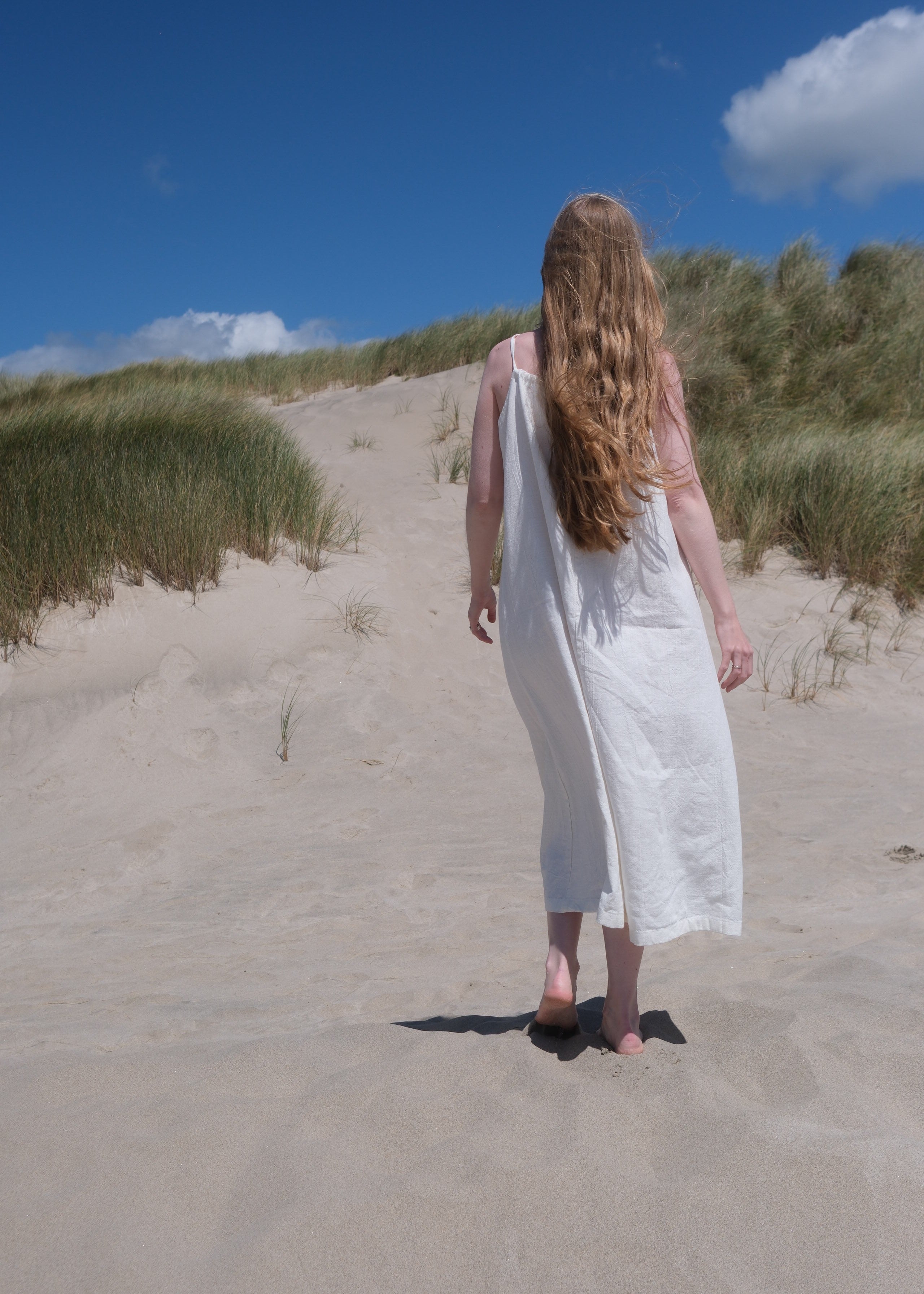 Person in a white dress standing on a sandy beach with grassy dunes and blue sky.