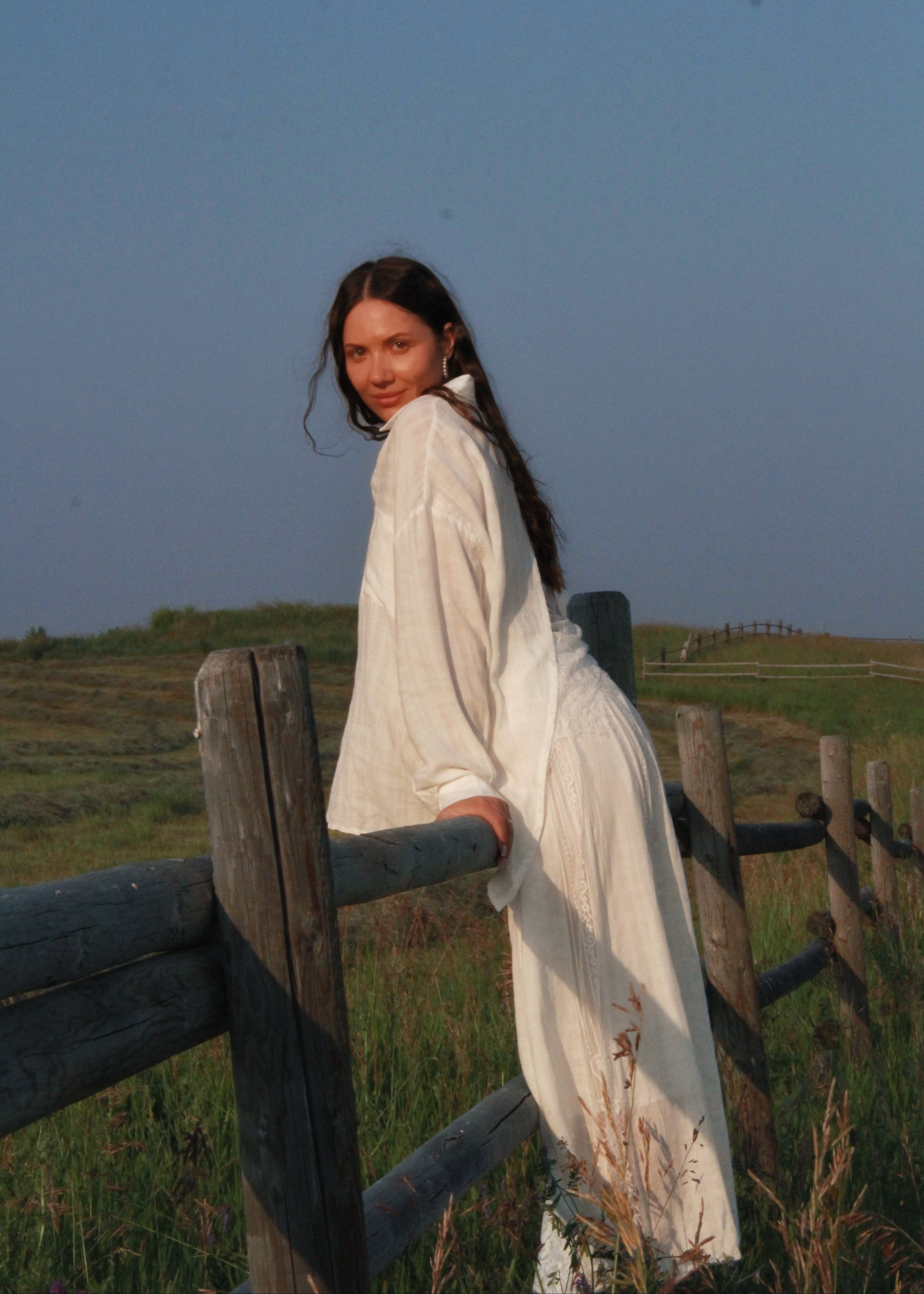 A woman in a long white dress leans against a rustic wooden fence in an open grassy field under a clear blue sky.
