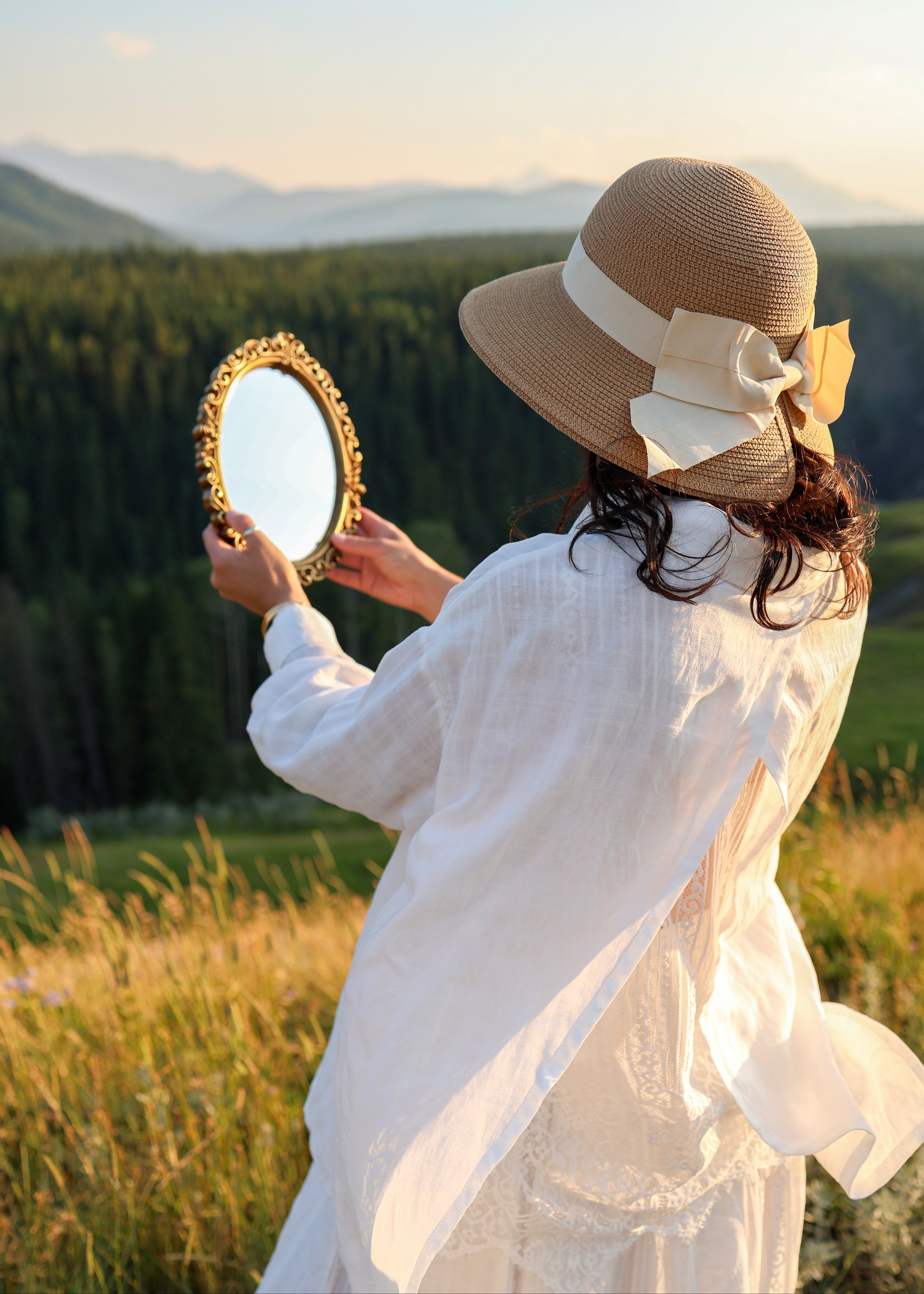 A woman in a white dress and a beige straw sun hat with a large bow stands outdoors in a grassy field, holding a small gold-framed mirror. Mountains and trees are visible in the background during golden hour.