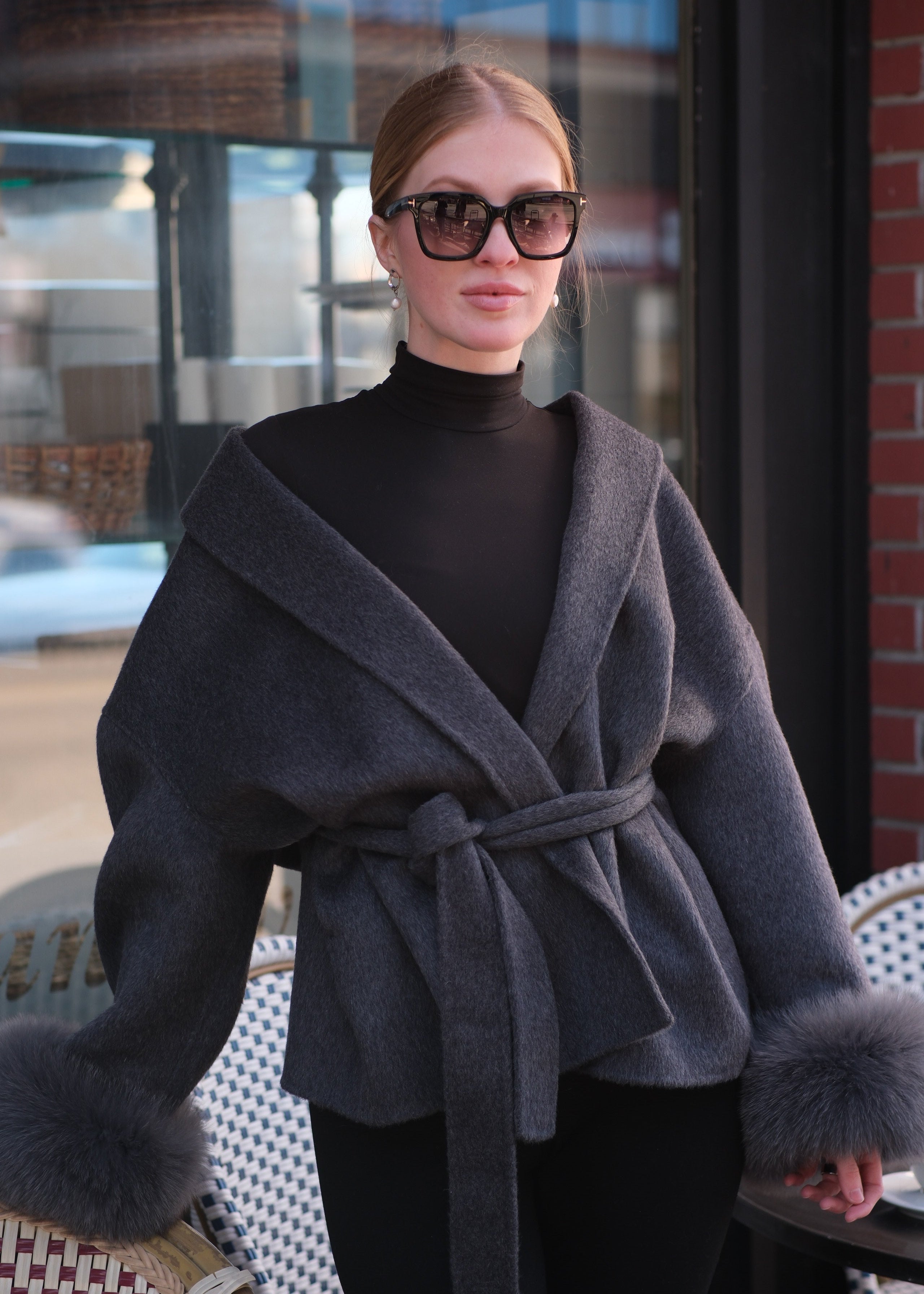 Person wearing a gray coat with a belt, standing in front of a store window.