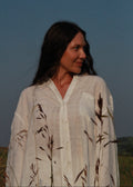 A woman wearing a loose white linen button-up shirt with dark brown grass motifs, standing outdoors against a blue sky.