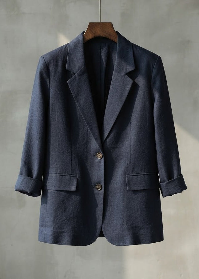 A dark navy blue linen blazer hanging on a wooden hanger against a textured gray wall.
