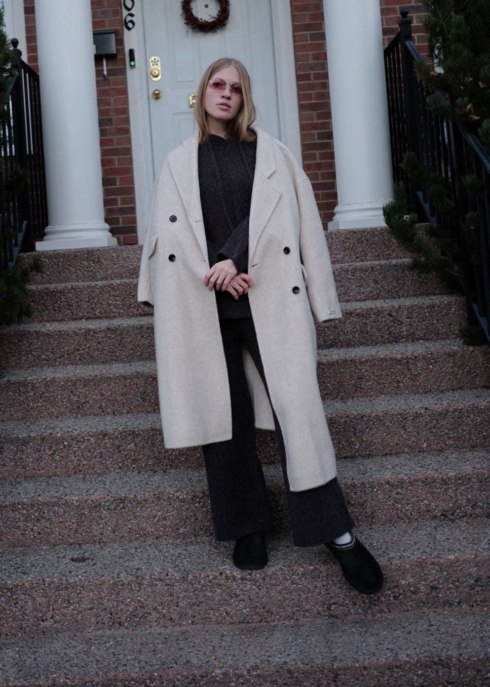 A woman stands on the front steps of a brick house wearing a long cream double-breasted coat over a black outfit.