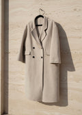 Beige coat hanging on a hanger against a light stone wall.