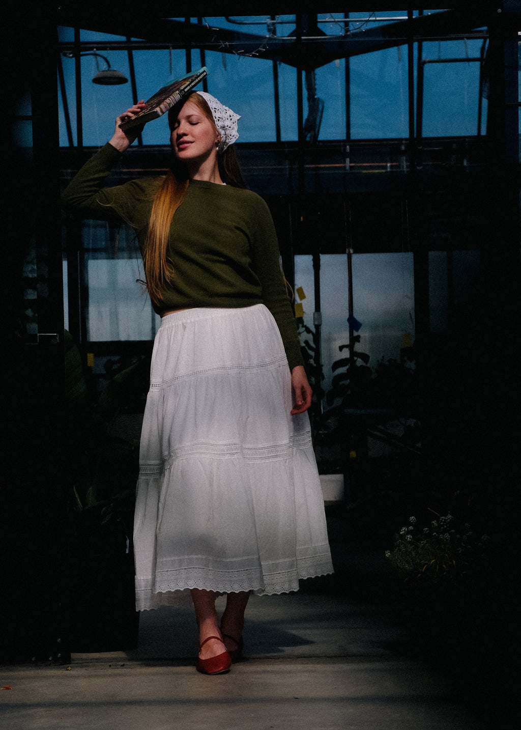 Woman in a green sweater and white skirt holding a book in a dark, industrial setting.