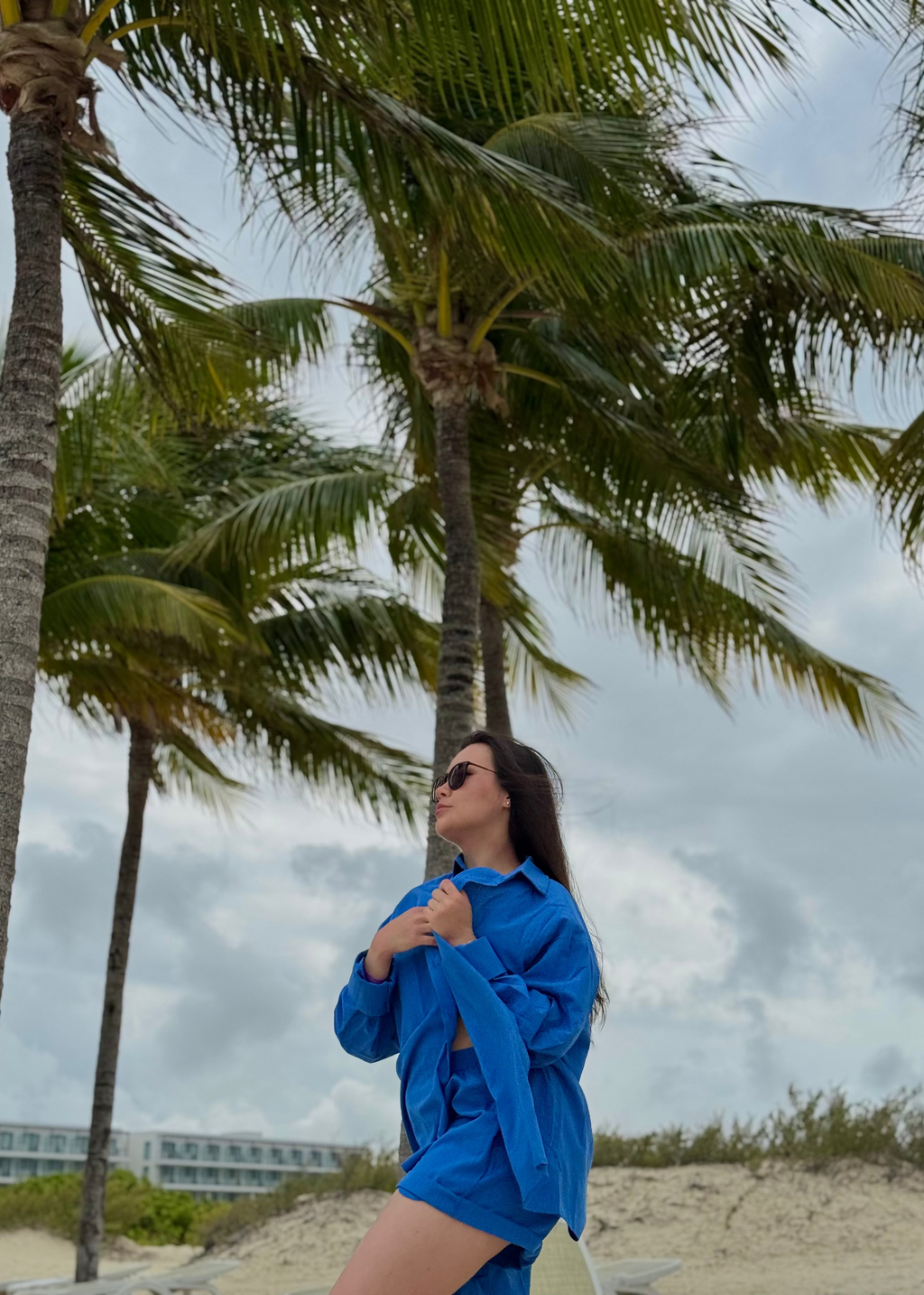 A woman wearing a bright blue oversized button-up shirt and matching shorts stands on a sandy beach with palm trees and a cloudy sky in the background.