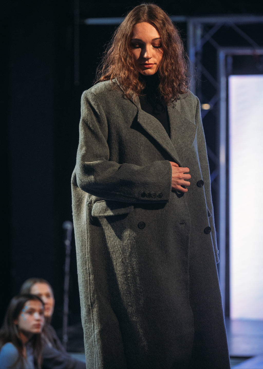 A model walks the runway wearing a long, double-breasted charcoal gray wool-blend coat over a black top. The photo is taken on a fashion show stage with a dark background and a bright screen in the distance.
