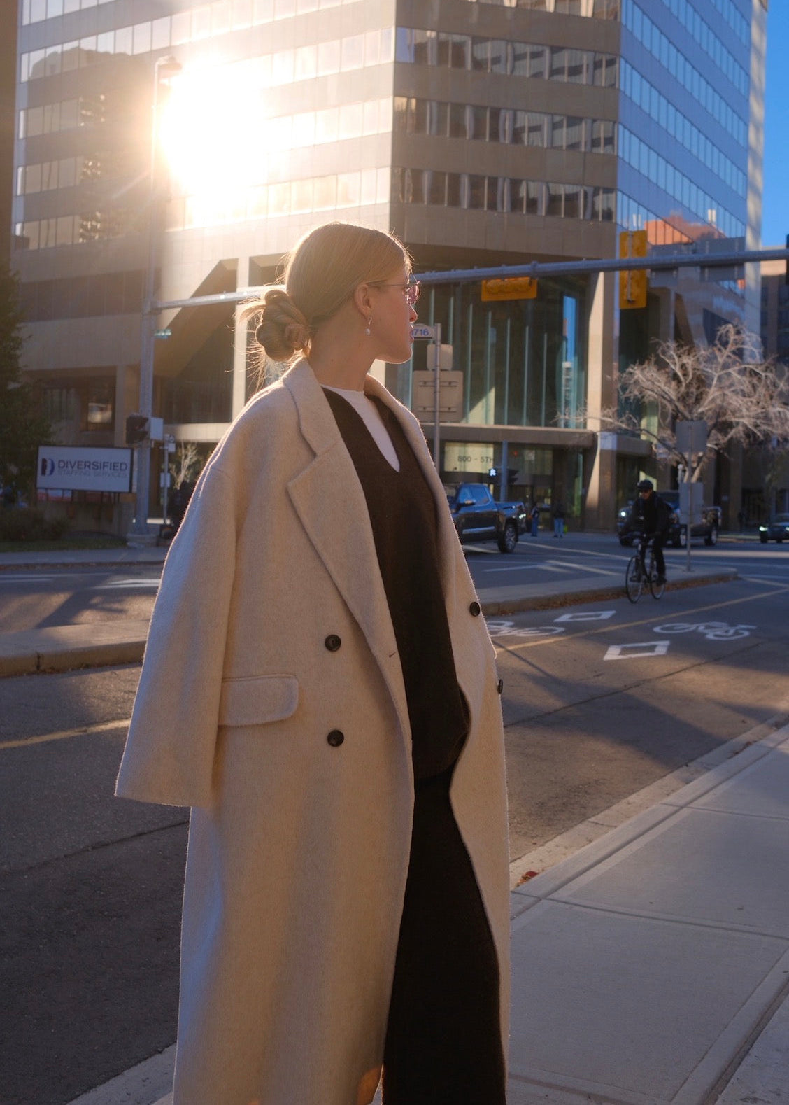 A woman wearing a long cream-colored double-breasted coat over a black dress stands on a city street corner with buildings and bright sunlight in the background.