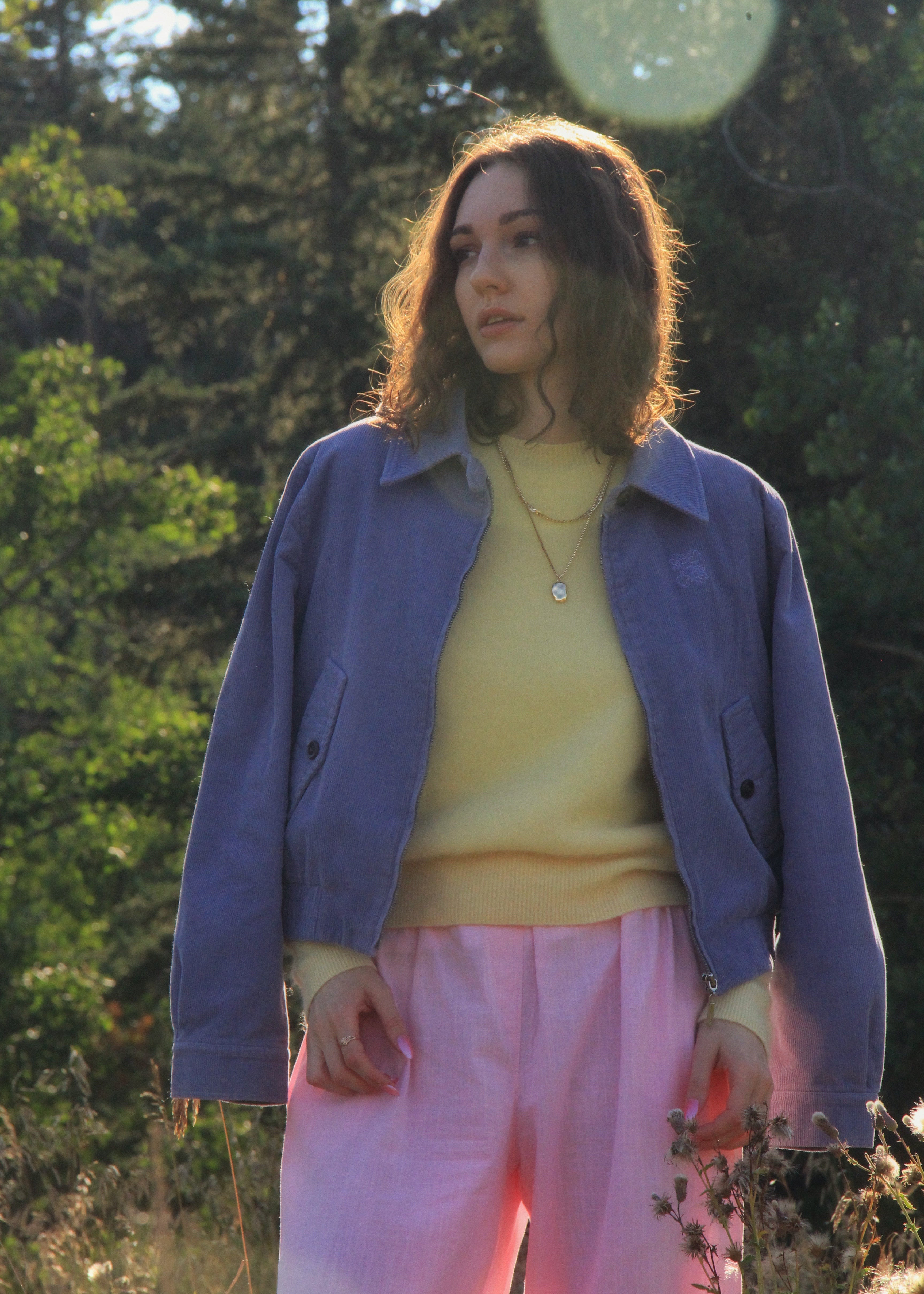 A woman standing outdoors in a wooded setting, wearing a lavender-blue jacket over a pale yellow top and pink pants.