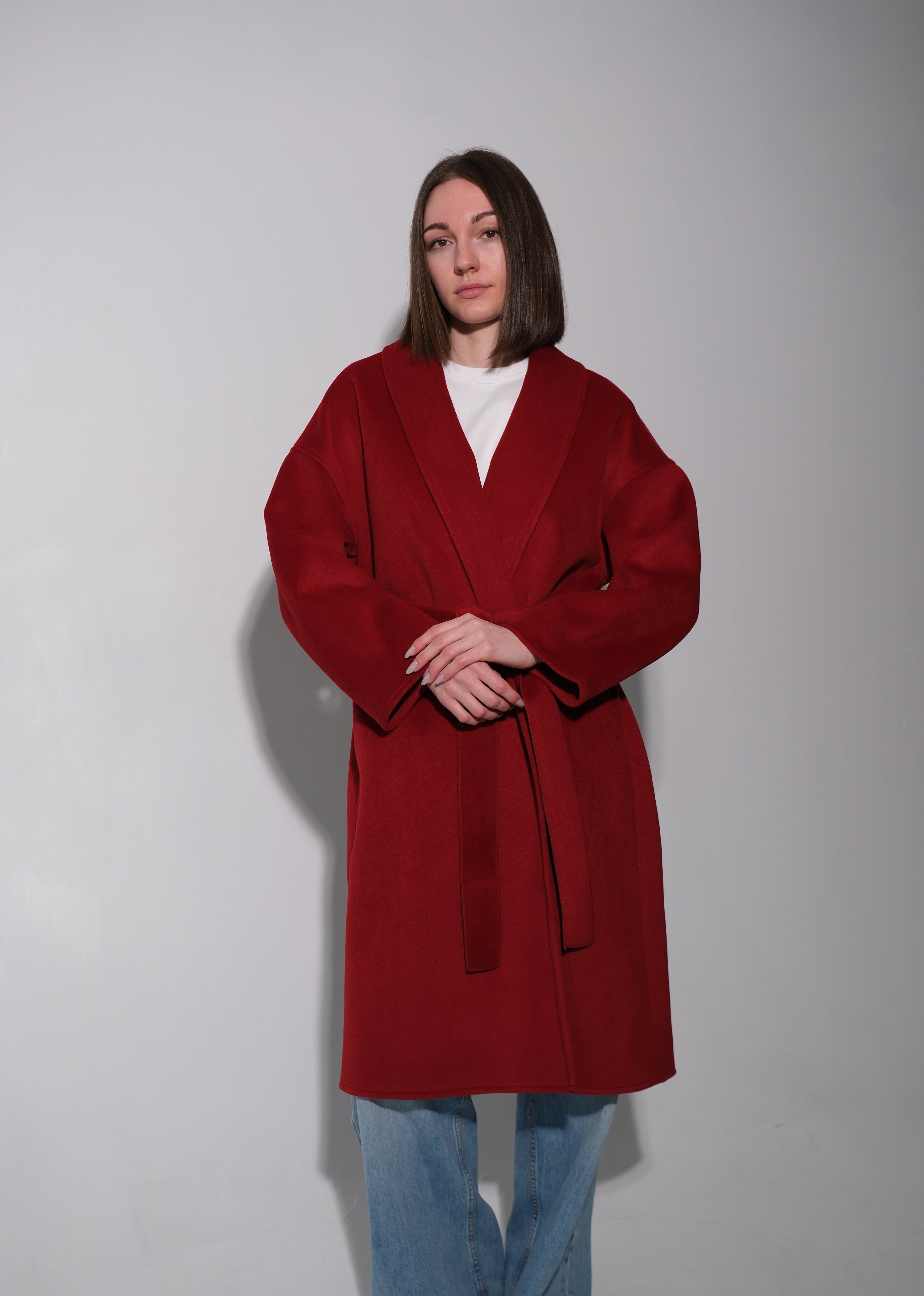 A woman modeling a long, oversized red wrap coat with a tie belt, worn over a white top and light blue jeans, standing against a plain light gray studio background.