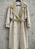 A beige trench coat with a contrasting tan suede collar and matching belt, hanging on a black hanger against a textured gray concrete wall.