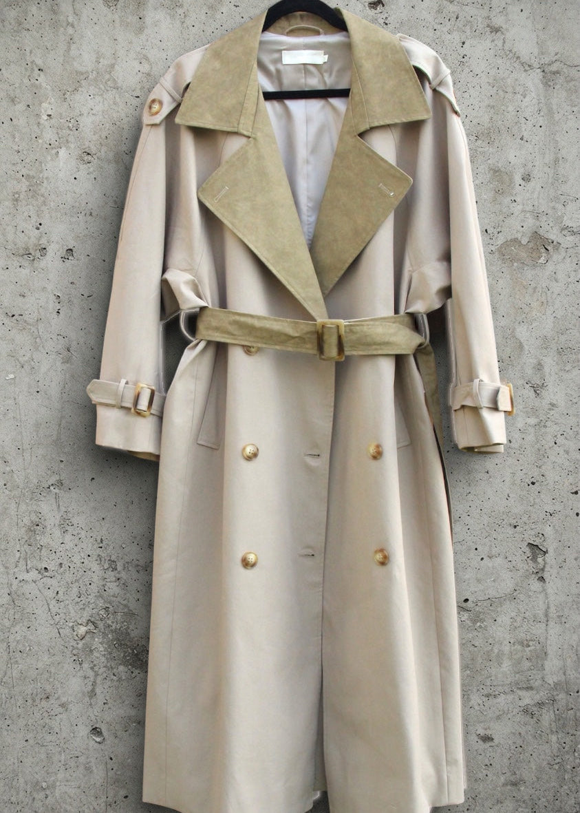 A beige trench coat with a contrasting tan suede collar and matching belt, hanging on a black hanger against a textured gray concrete wall.