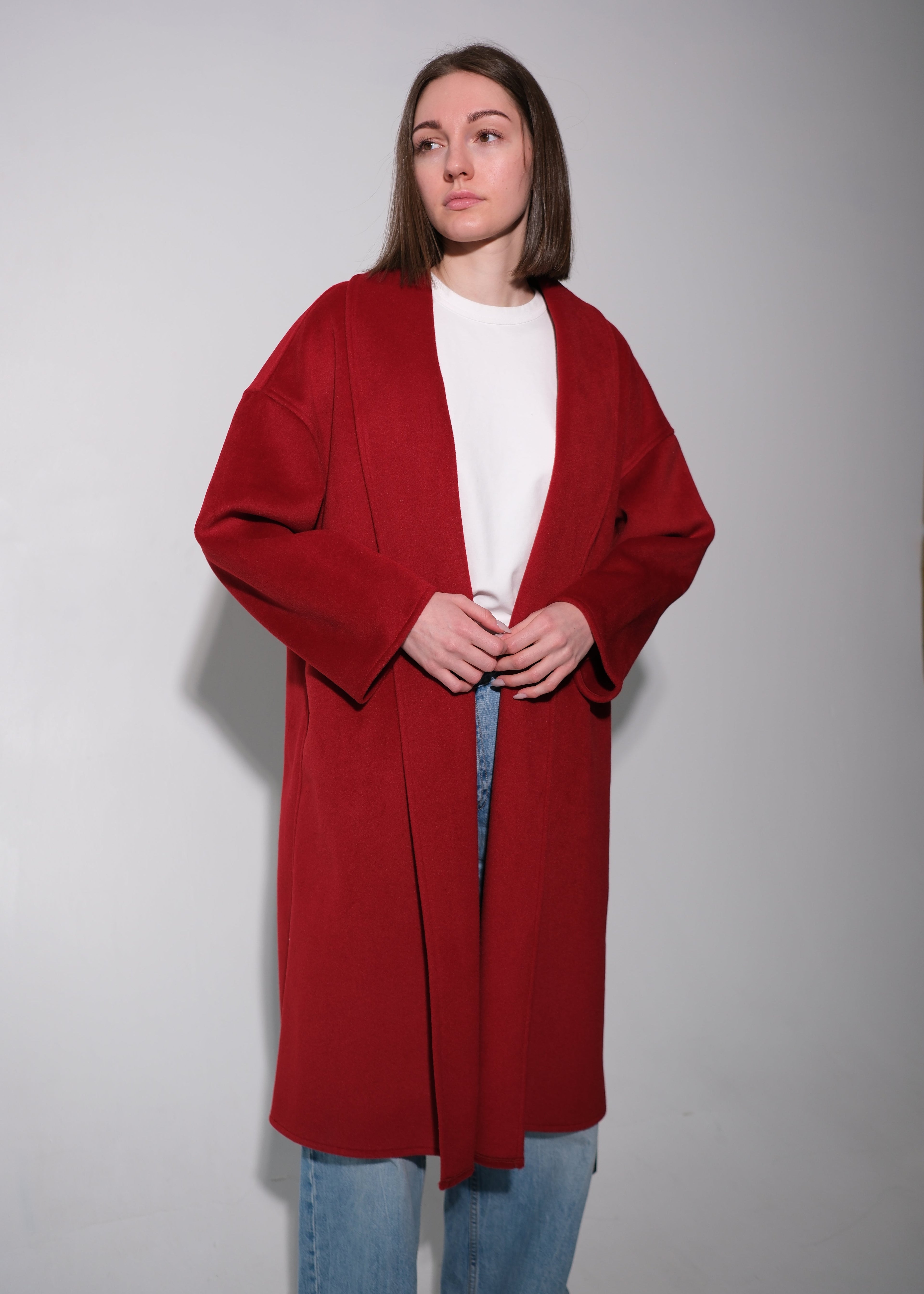 A woman modeling a long, open-front red cardigan over a white top and blue jeans, standing against a plain light gray studio background.