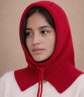 Person wearing a red knitted hood over a white garment against a beige background