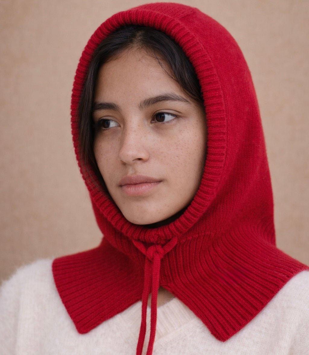 Person wearing a red knitted hood over a white garment against a beige background