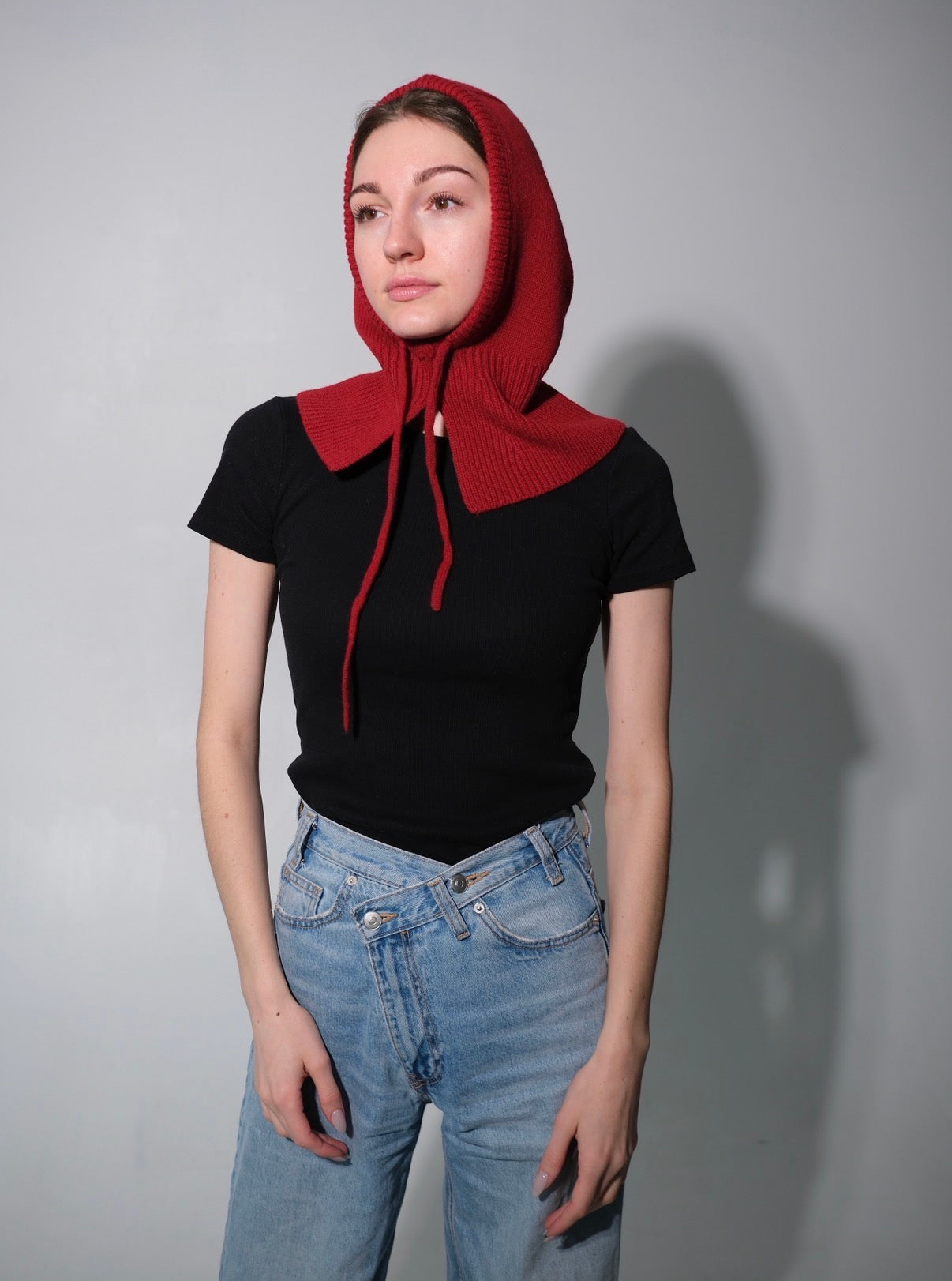 Person wearing a red hooded garment over a black top and blue jeans on a gray background. Wool balaclava for winter