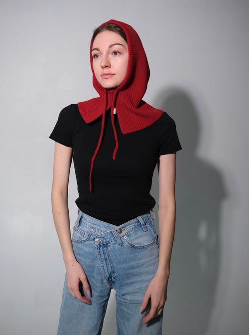 Person wearing a red hooded garment over a black top and blue jeans on a gray background. Wool balaclava for winter