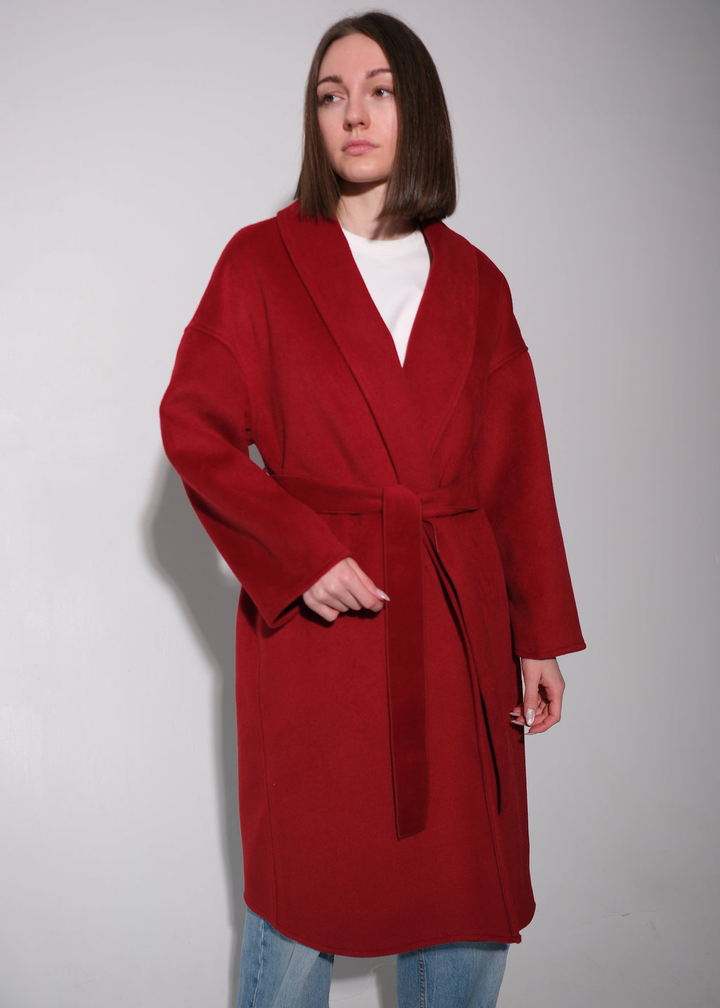 A woman wearing a long, deep red wrap-style coat with a tie belt over a white top and blue jeans, standing against a plain light gray wall.