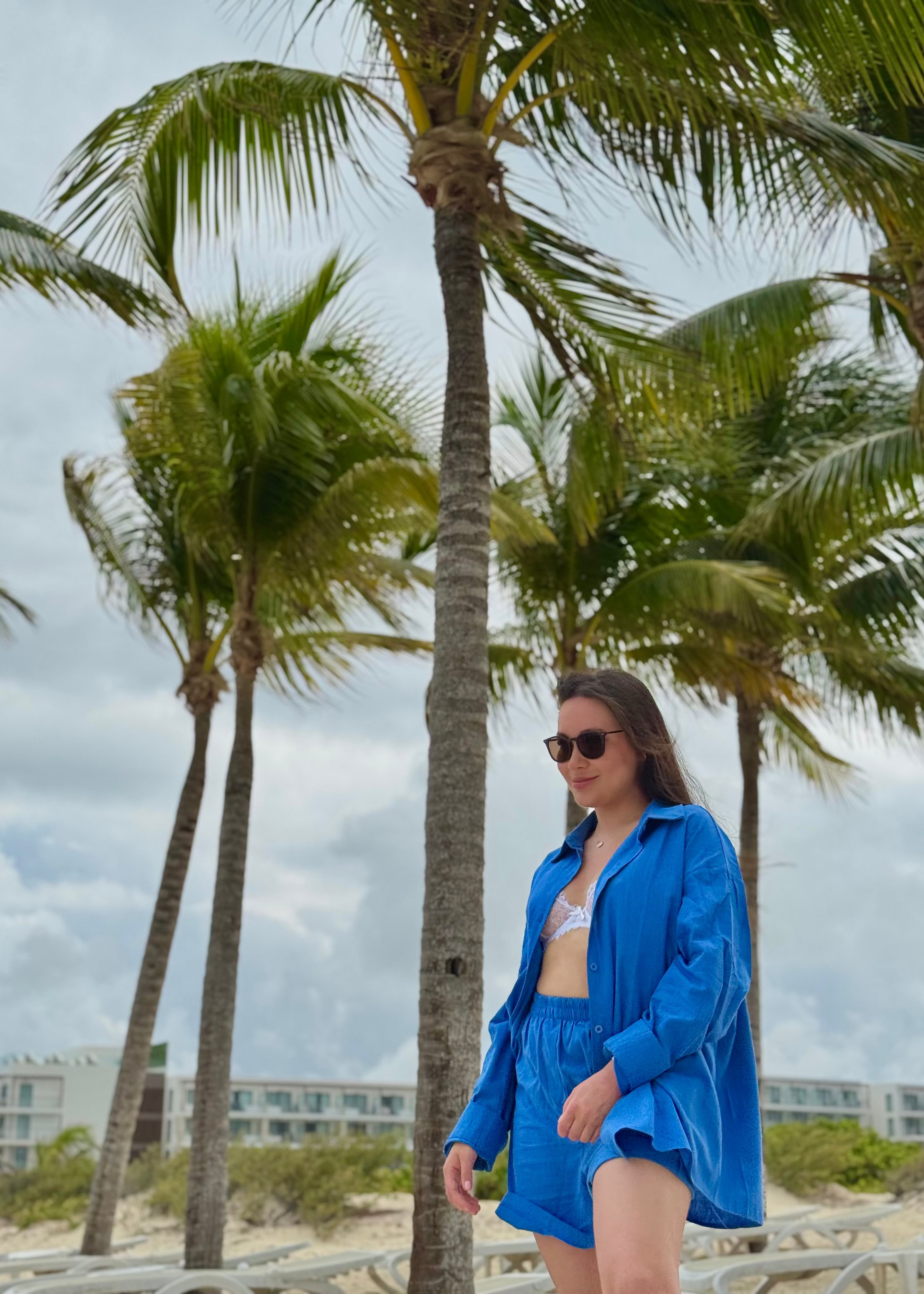 A woman wearing a bright blue matching lounge set and sunglasses stands outdoors on a beach walkway with palm trees and buildings in the background.