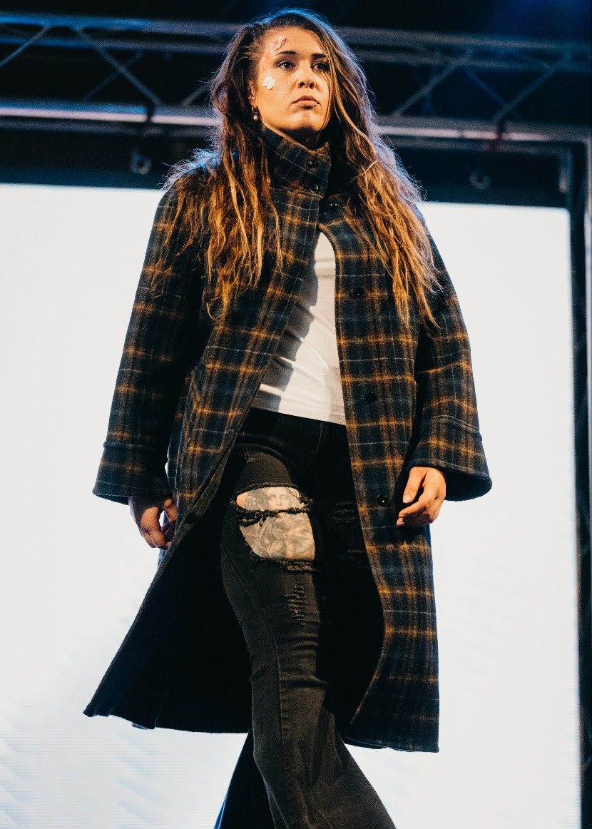 A woman modeling a long black and brown plaid coat over a white top and black distressed jeans with a ripped knee patch, standing on a runway.