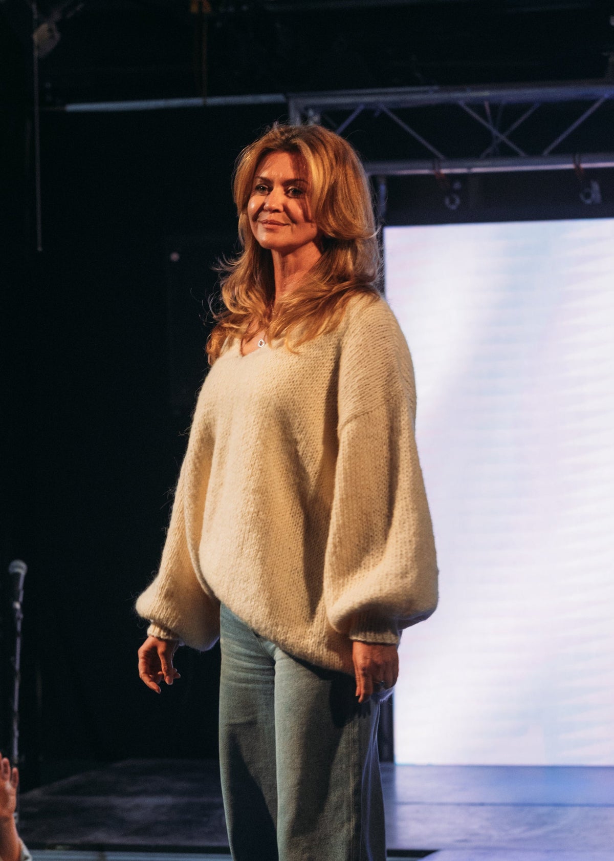 A woman stands on a runway wearing a cream-colored fuzzy knit sweater and wide-leg gray pants, with an audience seated in the foreground and a large screen behind her.