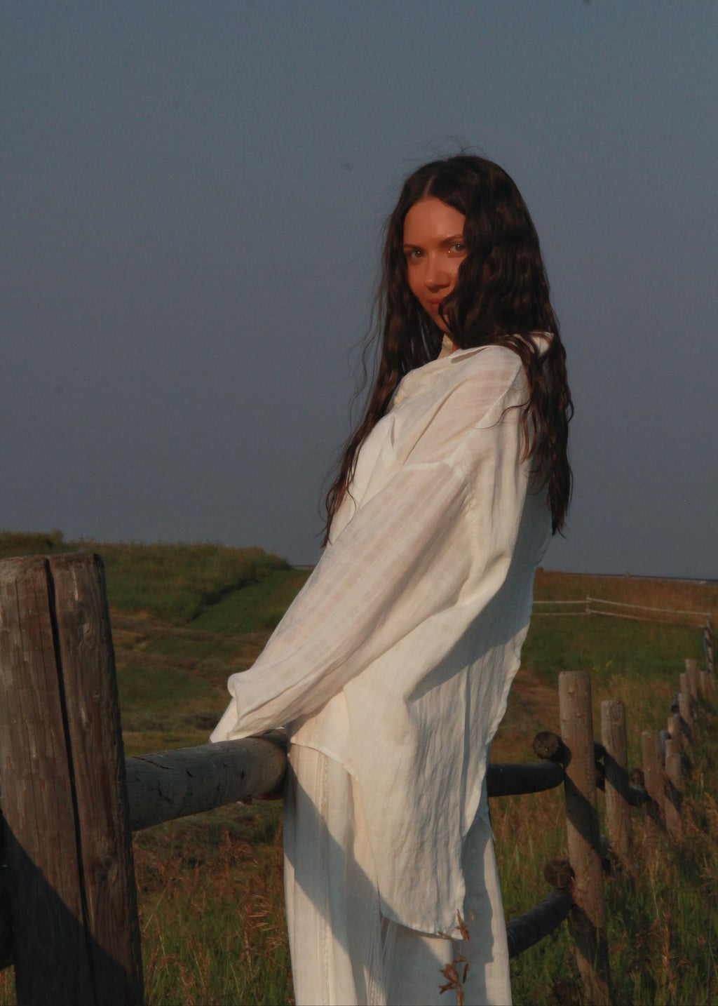 A woman with long dark hair stands outdoors near a wooden fence, wearing a loose white long-sleeve dress or robe in natural light.