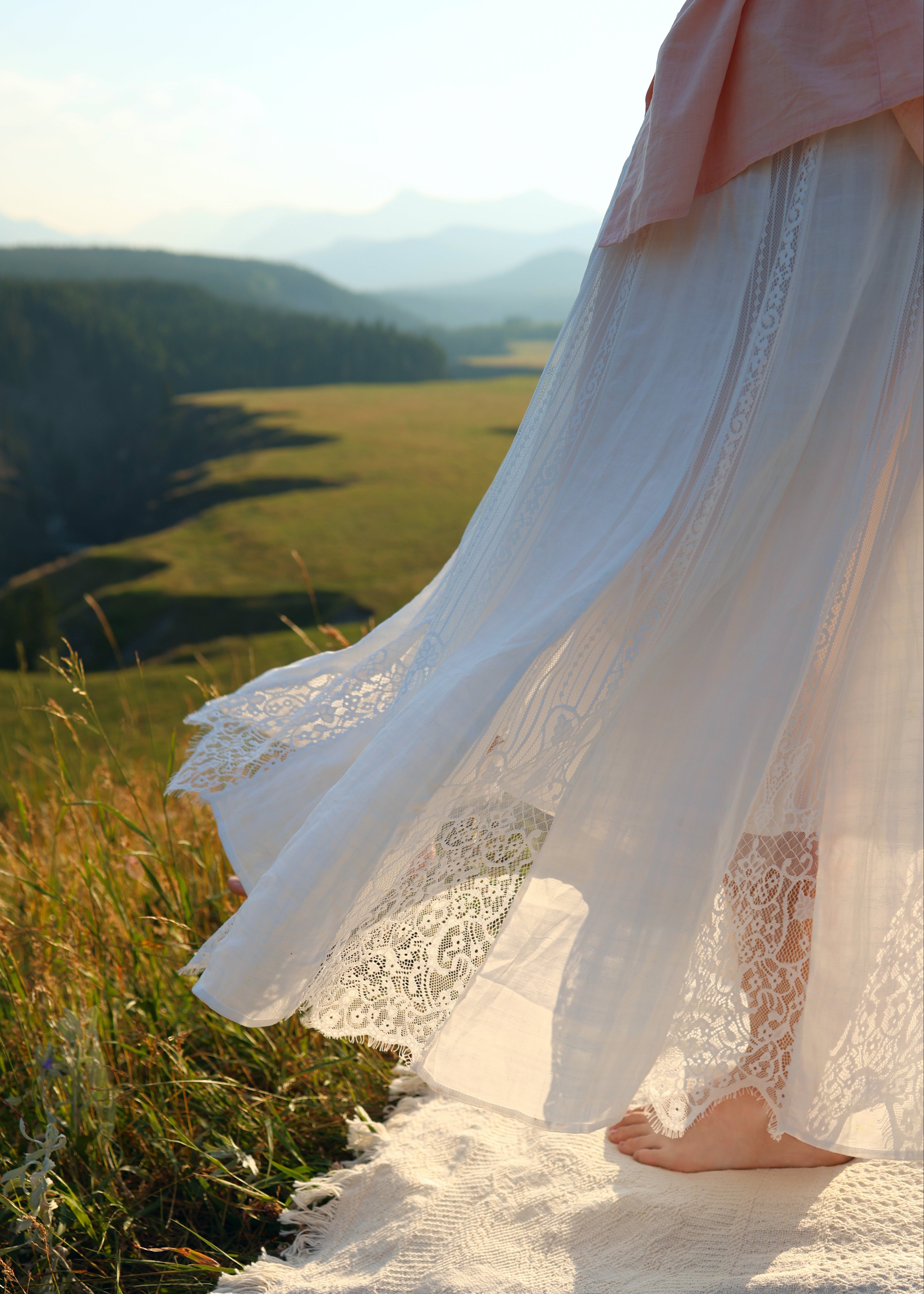 A flowing white maxi skirt with lace trim and a soft pink top, worn by a model standing barefoot on a white blanket in a sunlit mountain meadow.