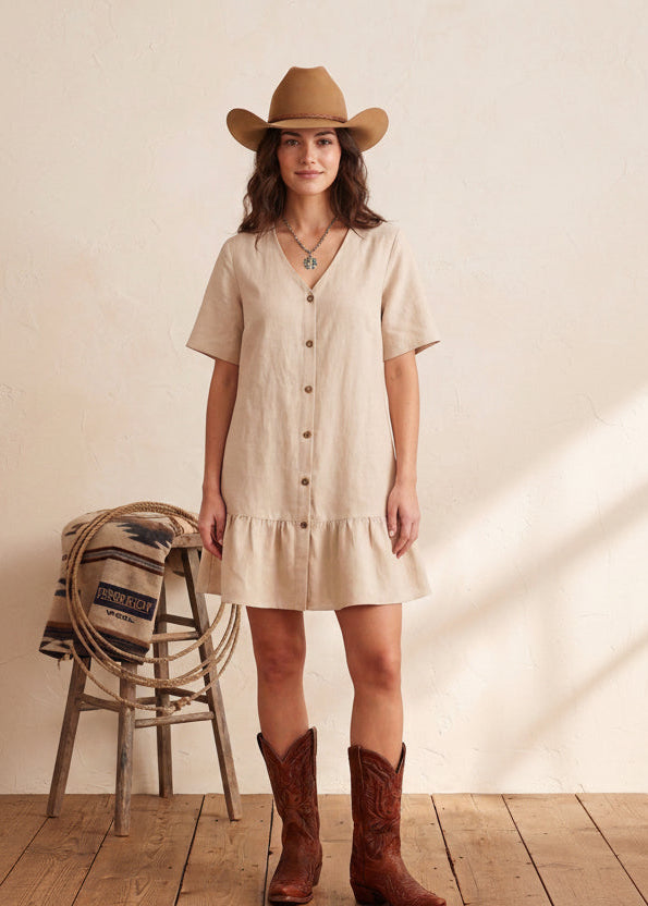 Woman wearing a beige dress, brown cowboy boots, and a cowboy hat in a rustic setting.