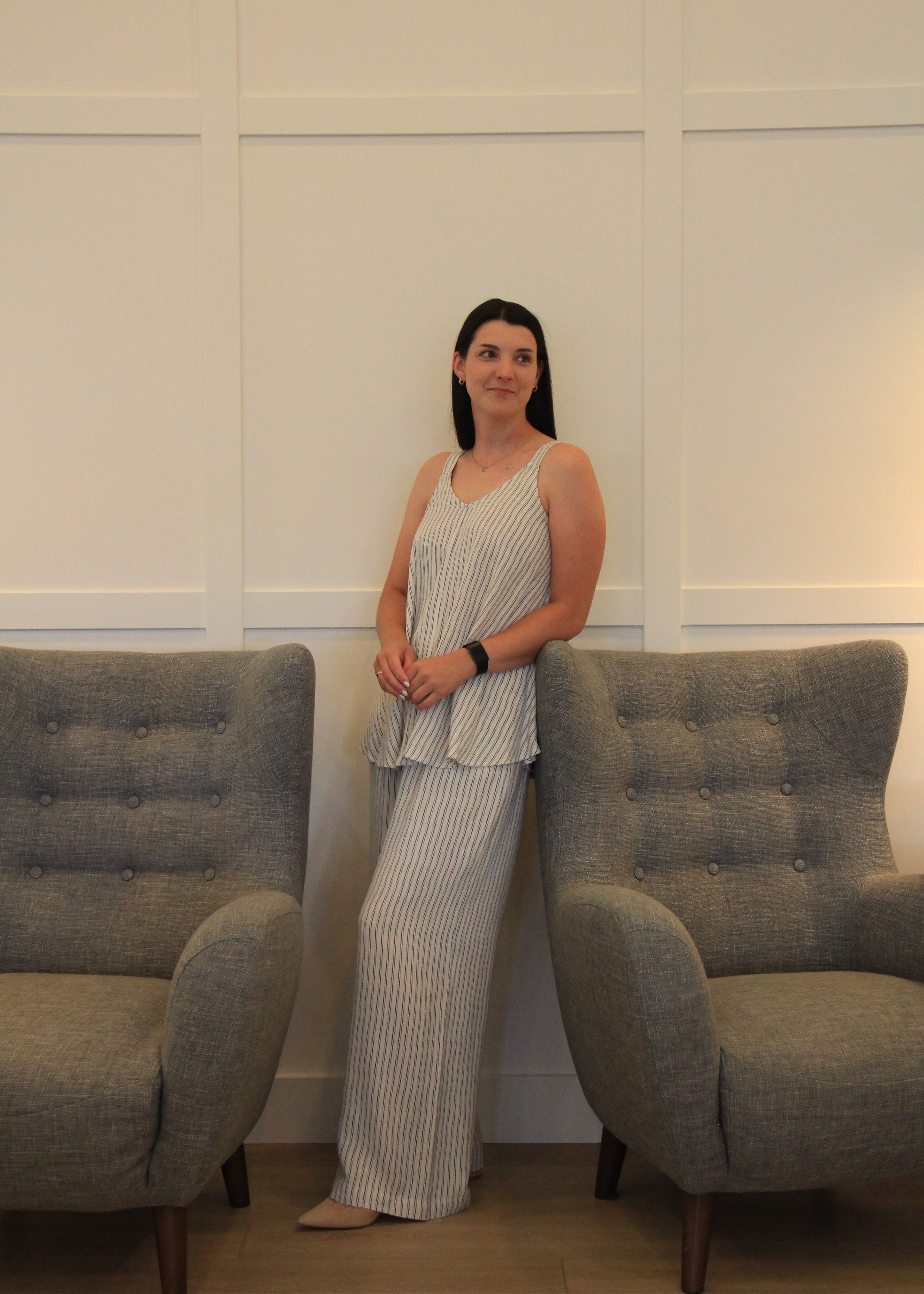 A woman stands between two gray tufted armchairs against a white paneled wall, wearing a sleeveless striped peplum top and matching wide-leg trousers in a light gray and white color-blocked set.