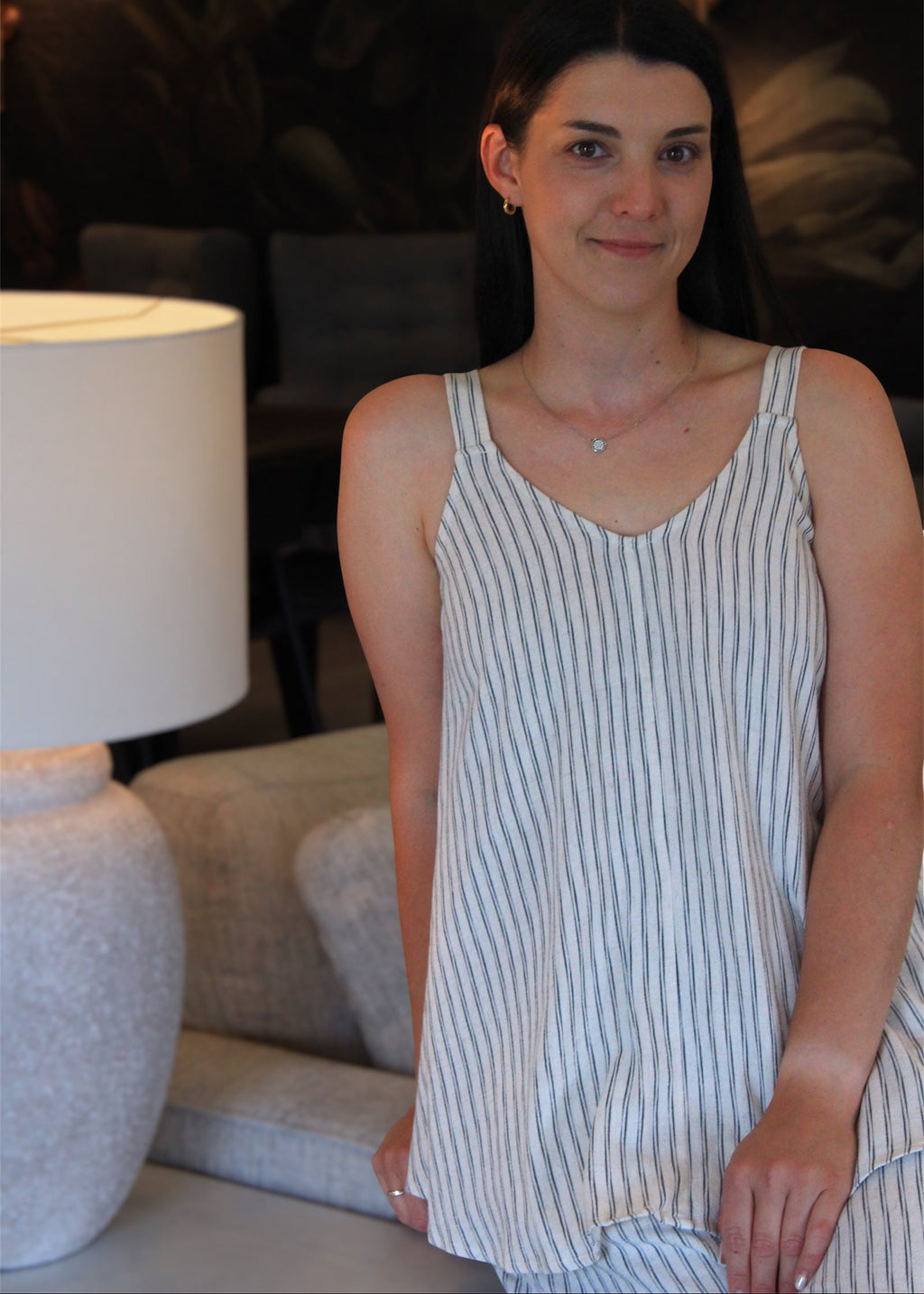 A woman wearing a sleeveless white camisole-style top with thin vertical navy stripes, standing indoors next to a lamp and sofa.