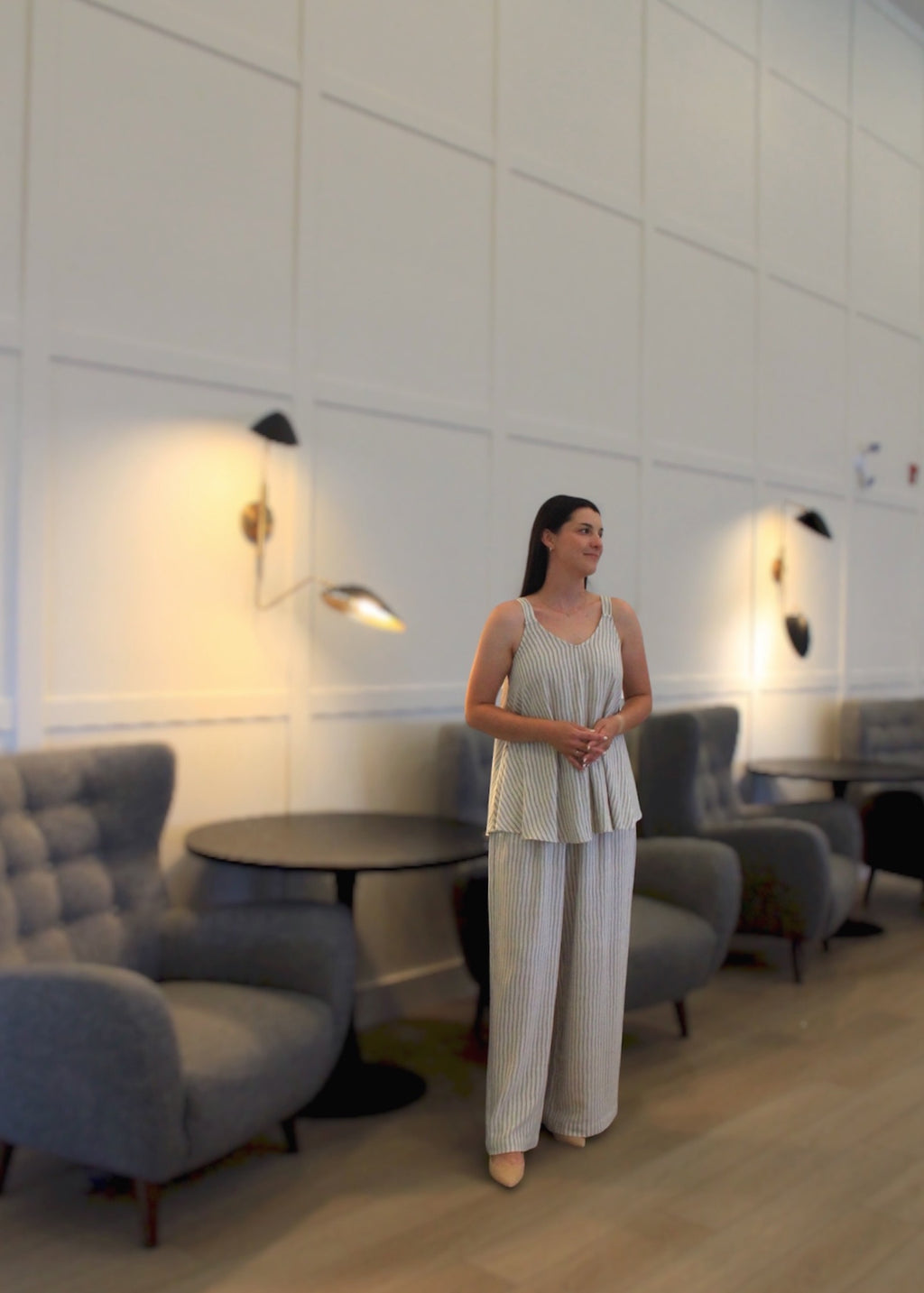 A woman wearing a light beige sleeveless peplum top and matching wide-leg trousers stands in a modern lounge area with gray tufted armchairs, round black tables, and warm wall sconces.