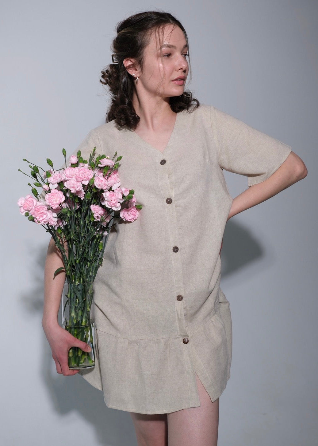 Woman holding a vase of pink flowers wearing a beige dress against a plain background