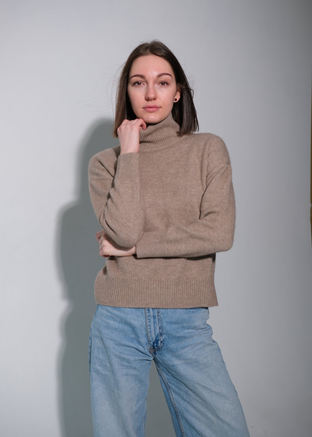 A woman wearing a light brown ribbed turtleneck sweater and light blue jeans, standing against a plain gray studio background.