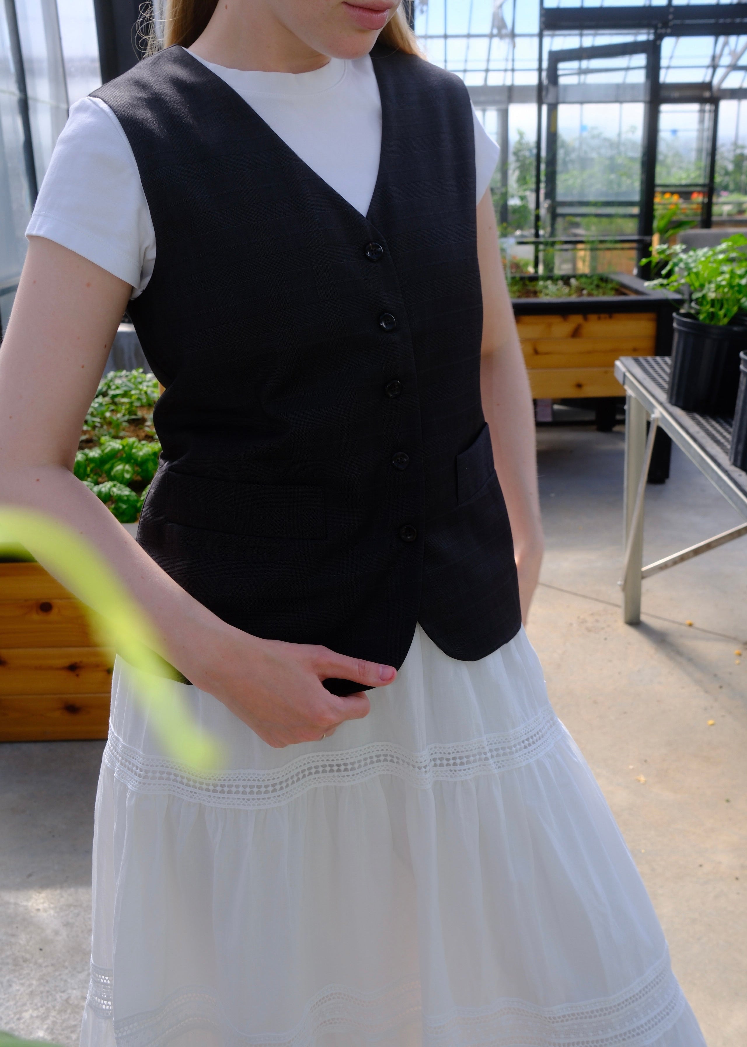 A woman wearing a black button-front vest over a white short-sleeve top and a long white skirt, standing in a greenhouse with plants and glass panels in the background.
