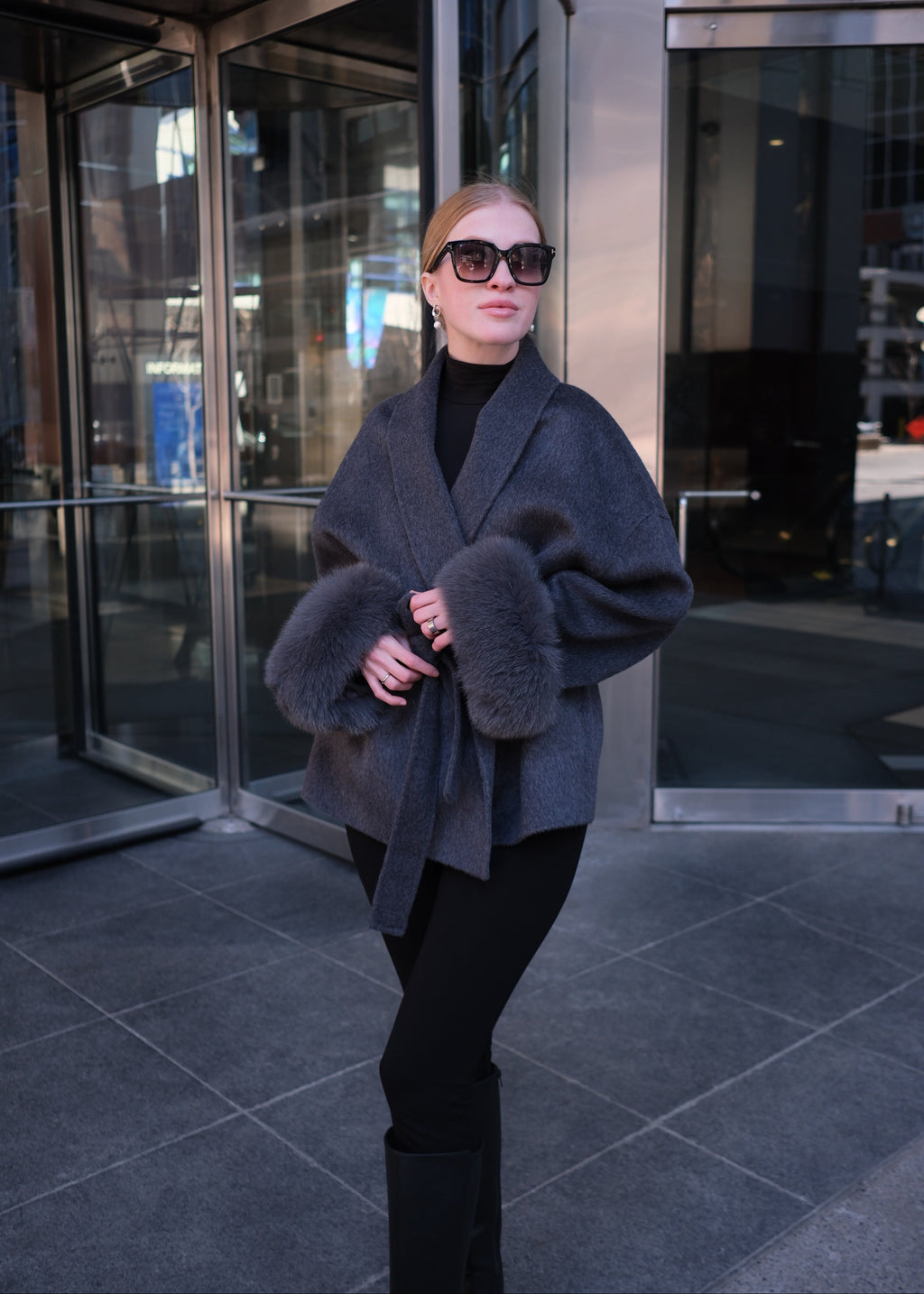 Person wearing a dark coat with fur trim standing in front of a reflective surface.