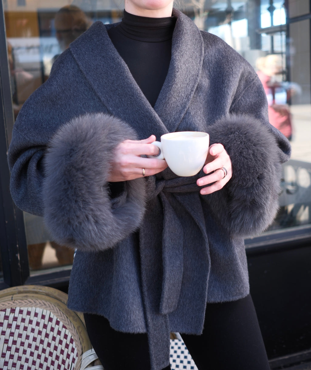 Person wearing a gray coat with fur cuffs holding a white mug.