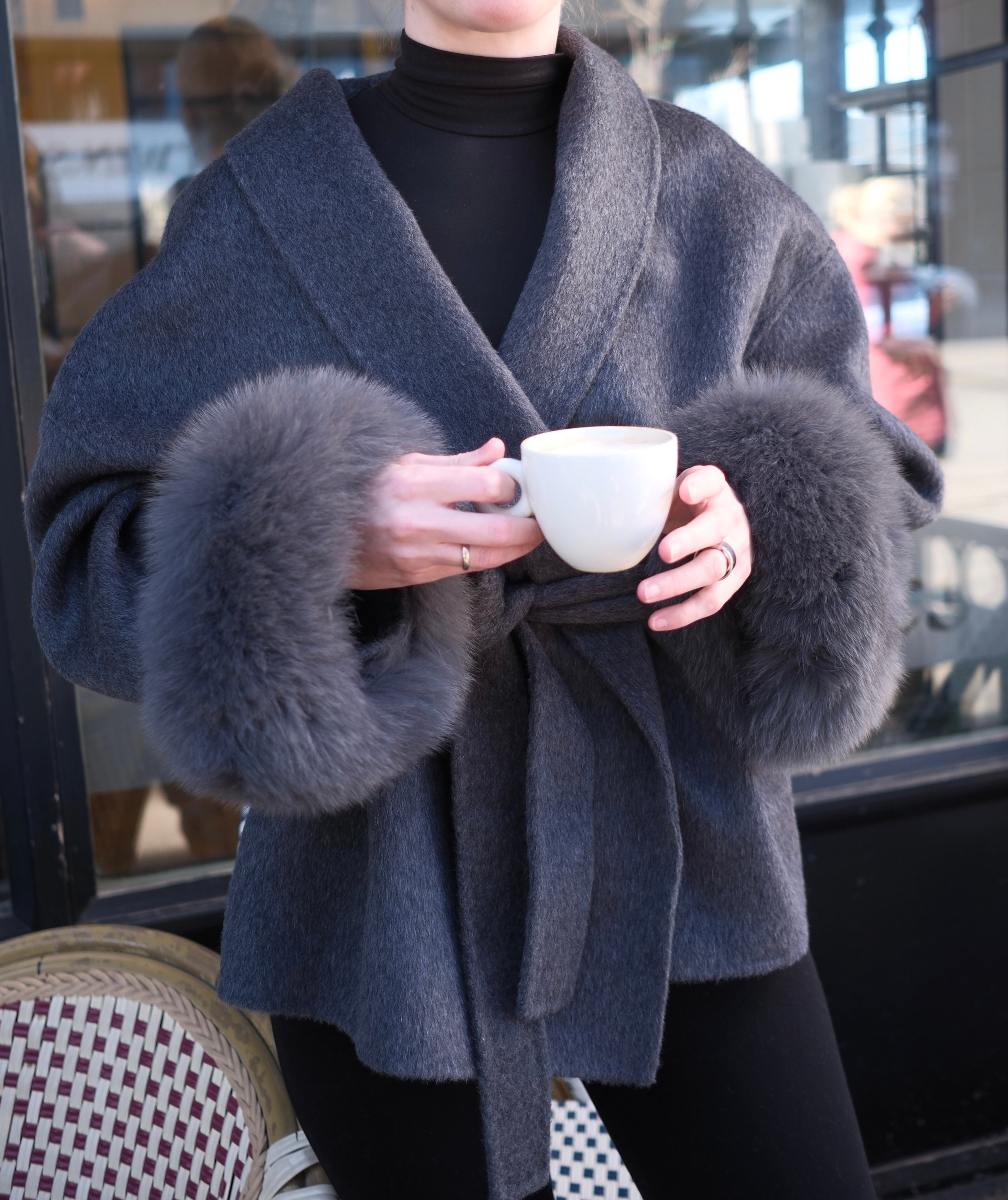 Person wearing a gray coat with fur cuffs holding a white mug.