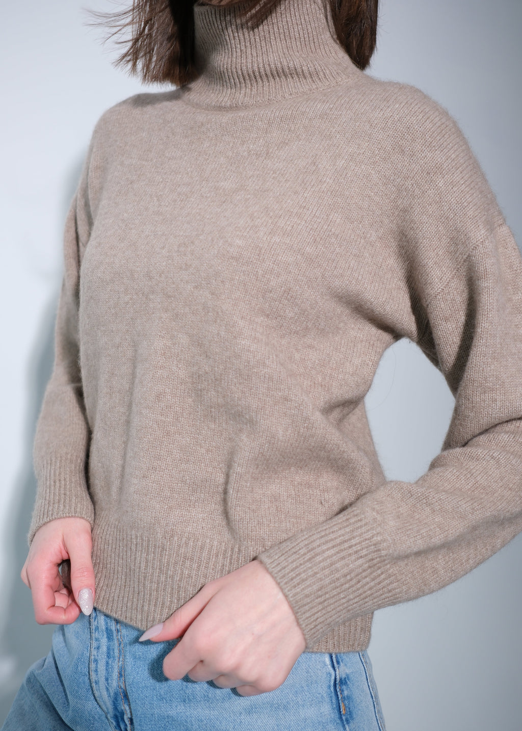 A person wearing a taupe beige knit turtleneck sweater with light blue jeans, shown from the neck down against a plain light gray background.