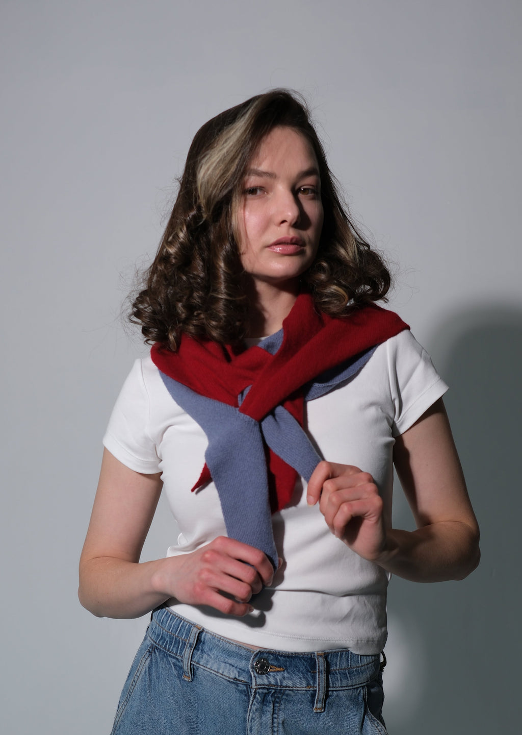 A woman with shoulder-length brown hair models a red and blue knit scarf tied around her neck, paired with a white t-shirt and blue jeans, against a plain gray studio background.