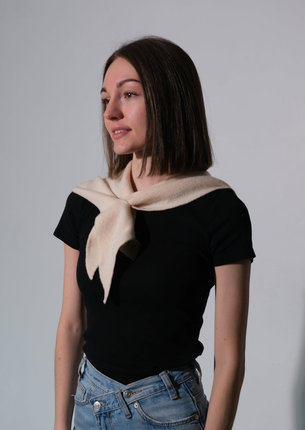 A woman wearing a black short-sleeve t-shirt, light-wash blue jeans, and a cream-colored knit scarf tied around her neck, standing against a plain light gray studio background.
