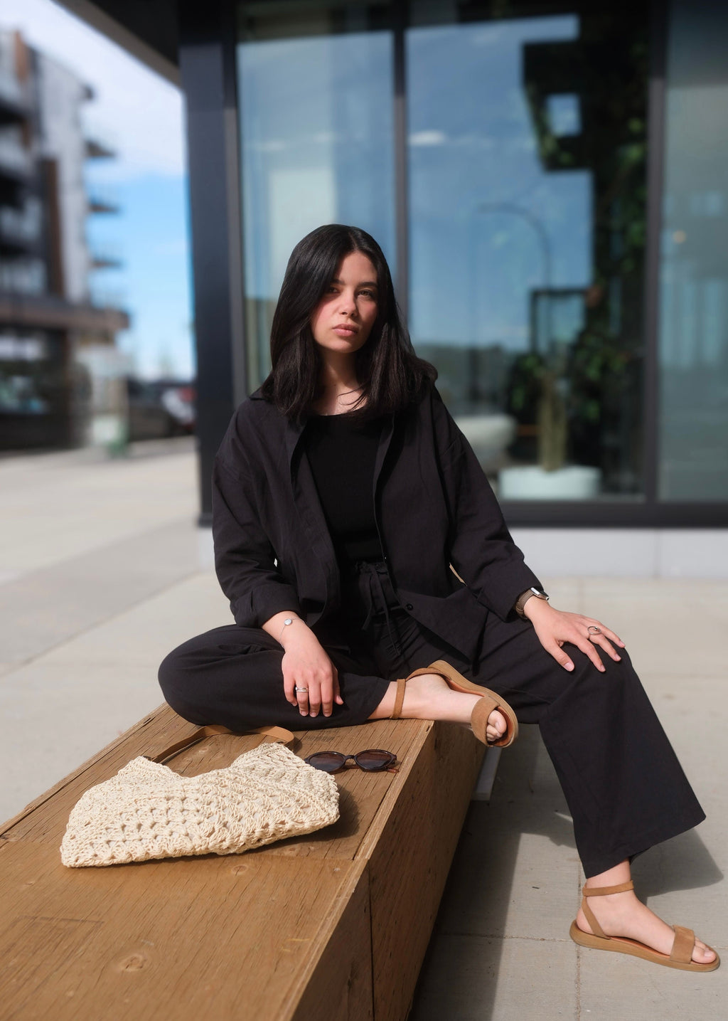 Woman in black outfit sitting on a wooden bench with a blurred urban background