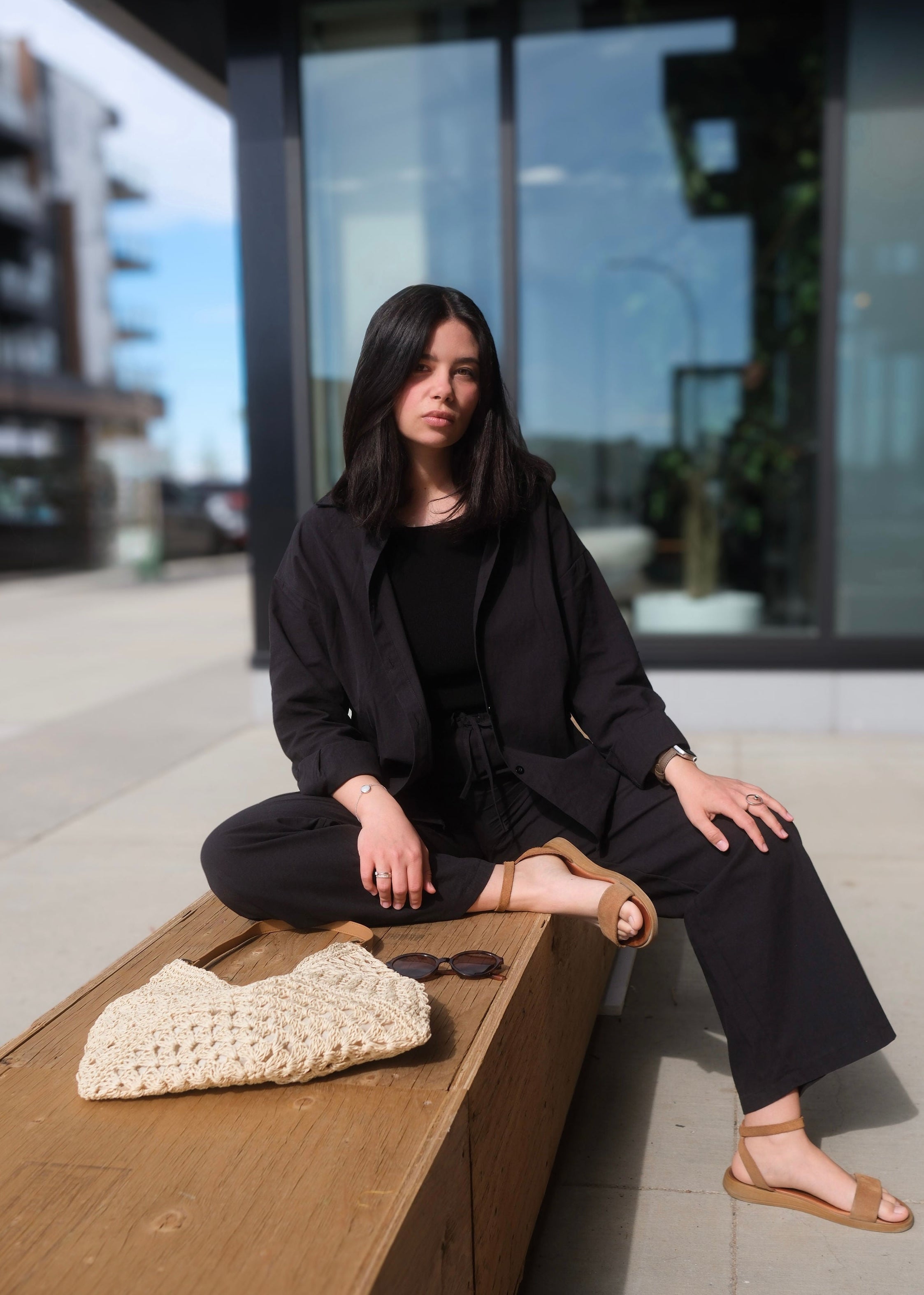 Woman in black outfit sitting on a wooden bench with a blurred urban background