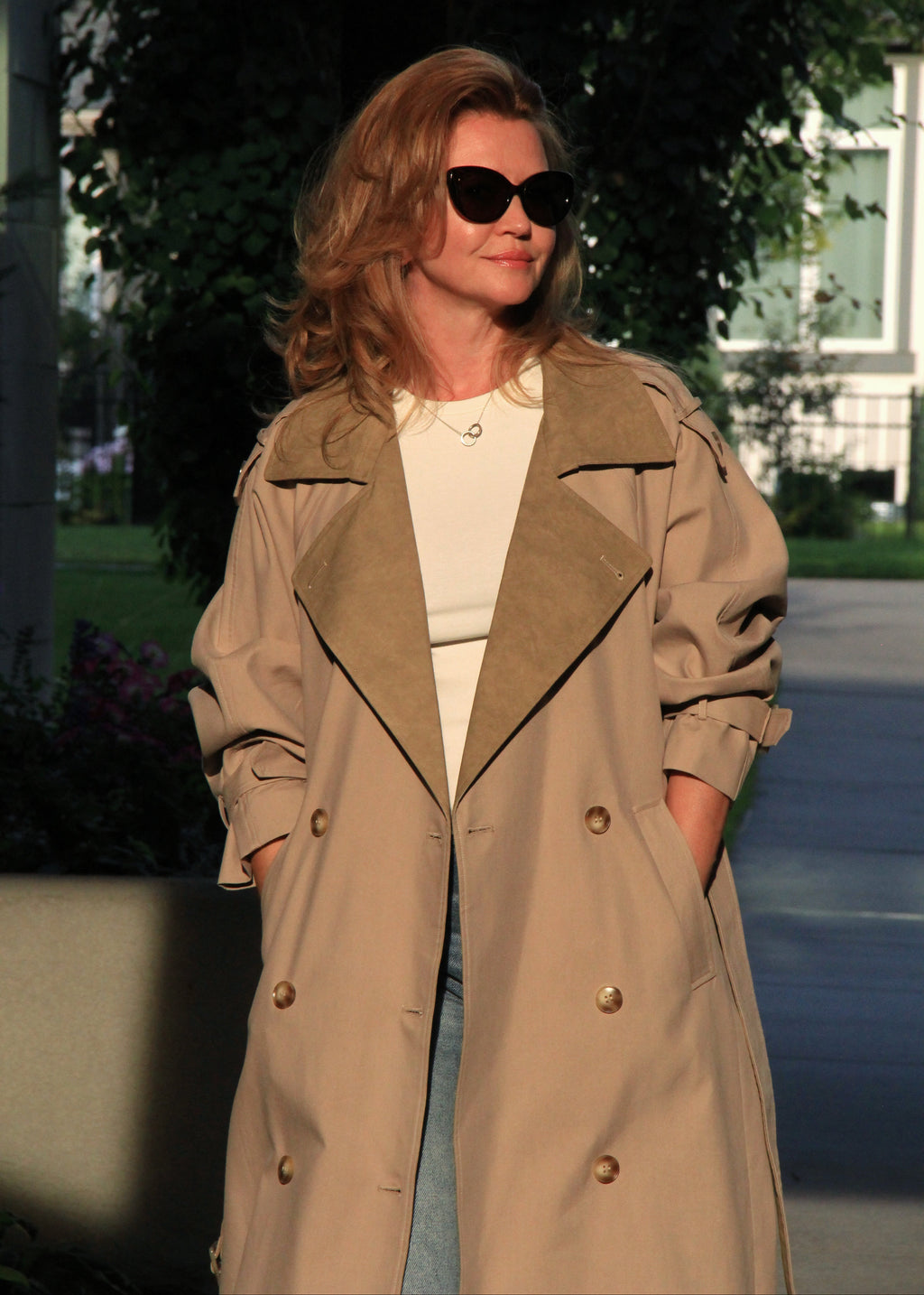 A woman wearing a beige double-breasted trench coat over a white top and light blue jeans, styled with black sunglasses, standing outdoors in front of a blurred background of trees and a building.